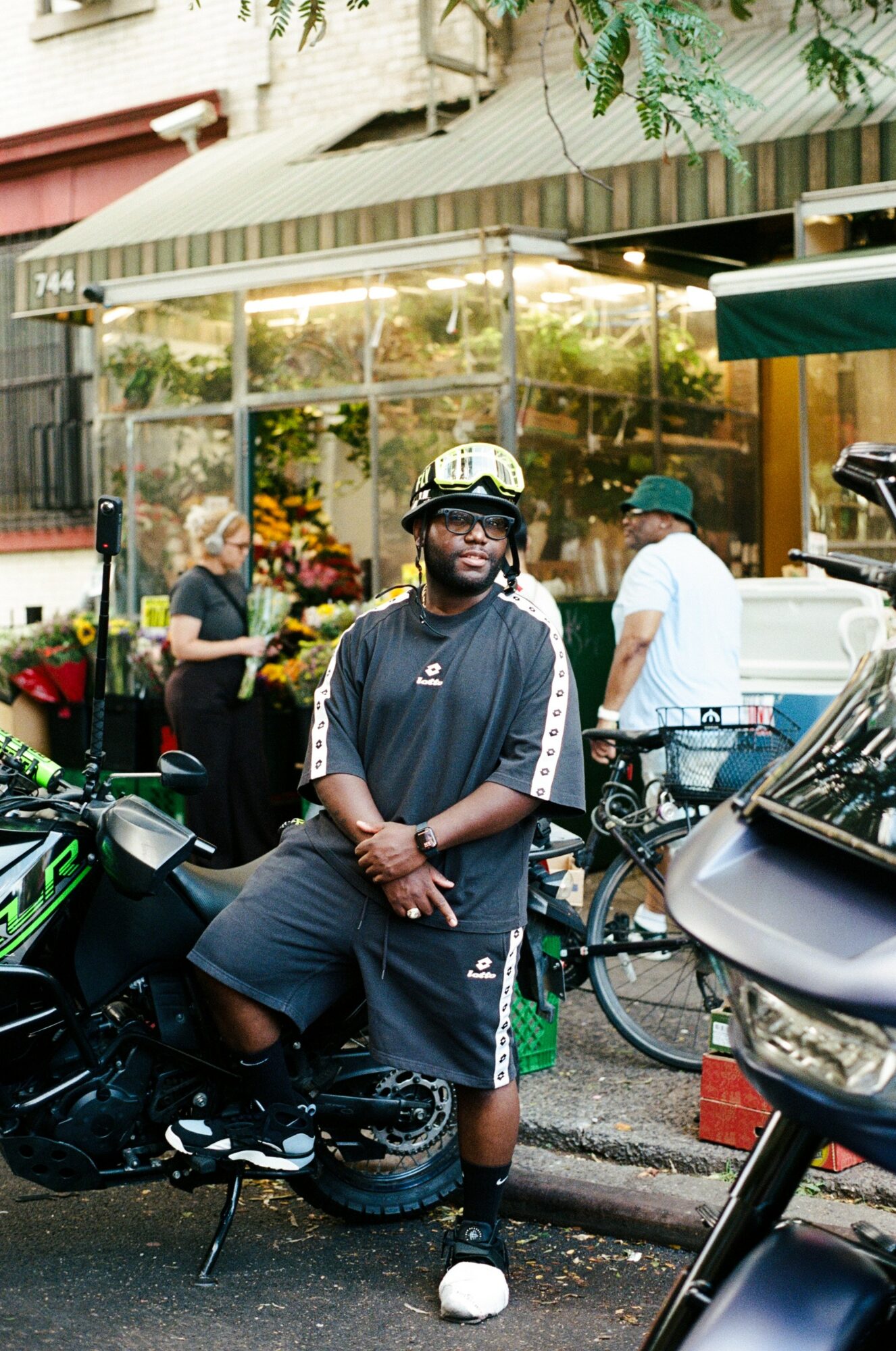 Man in sportswear standing next to a motorcycle on a city sidewalk, storefront with flowers in background.