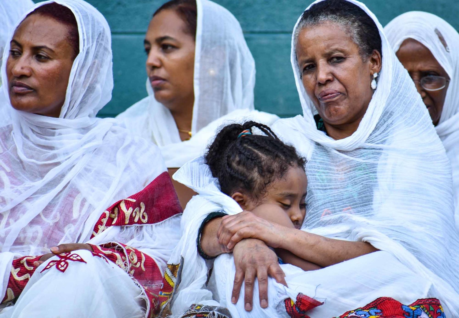 Group of women and a child, some wearing white head coverings, sitting together outdoors, one woman holding a child. Word partially visible on clothing.