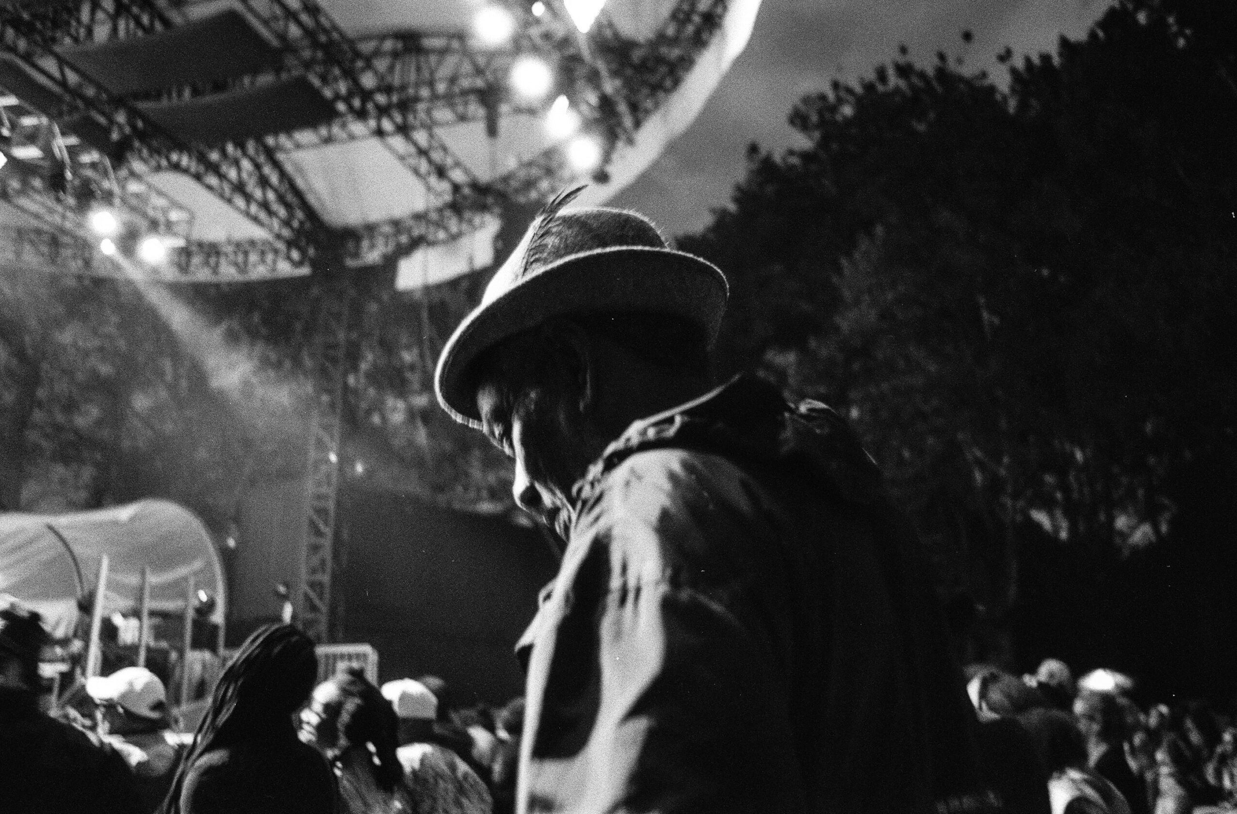 Person wearing a hat at an outdoor concert or event, with stage lights and crowd in background.