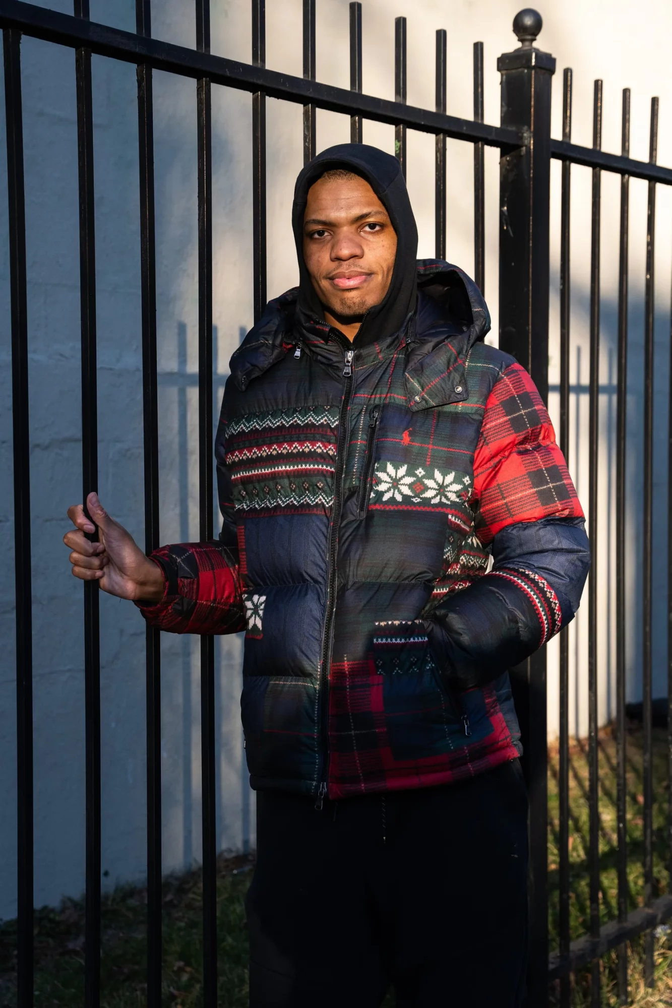 Young man standing outdoors near a black metal fence, wearing a colorful patterned jacket and black beanie, smiling.