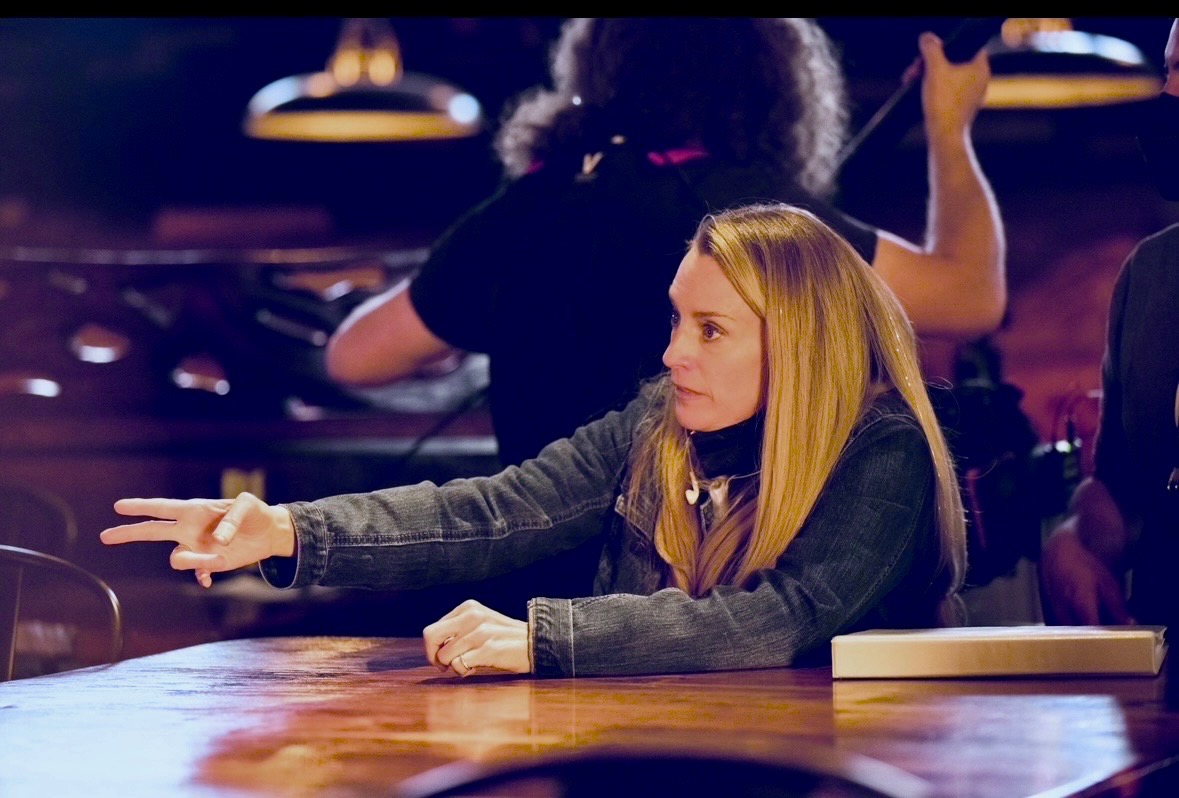 A woman with long blonde hair sits at a bar, reaching out with her right hand, with a person behind her in a black shirt. Word count: 20