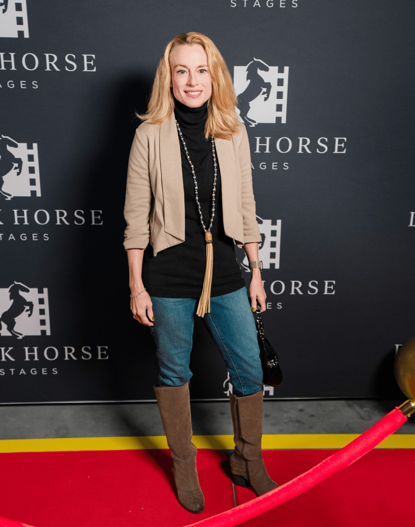Woman with blonde hair smiling, standing in front of a black backdrop with white text and logos, wearing a beige jacket, black top, blue jeans, and brown boots.
