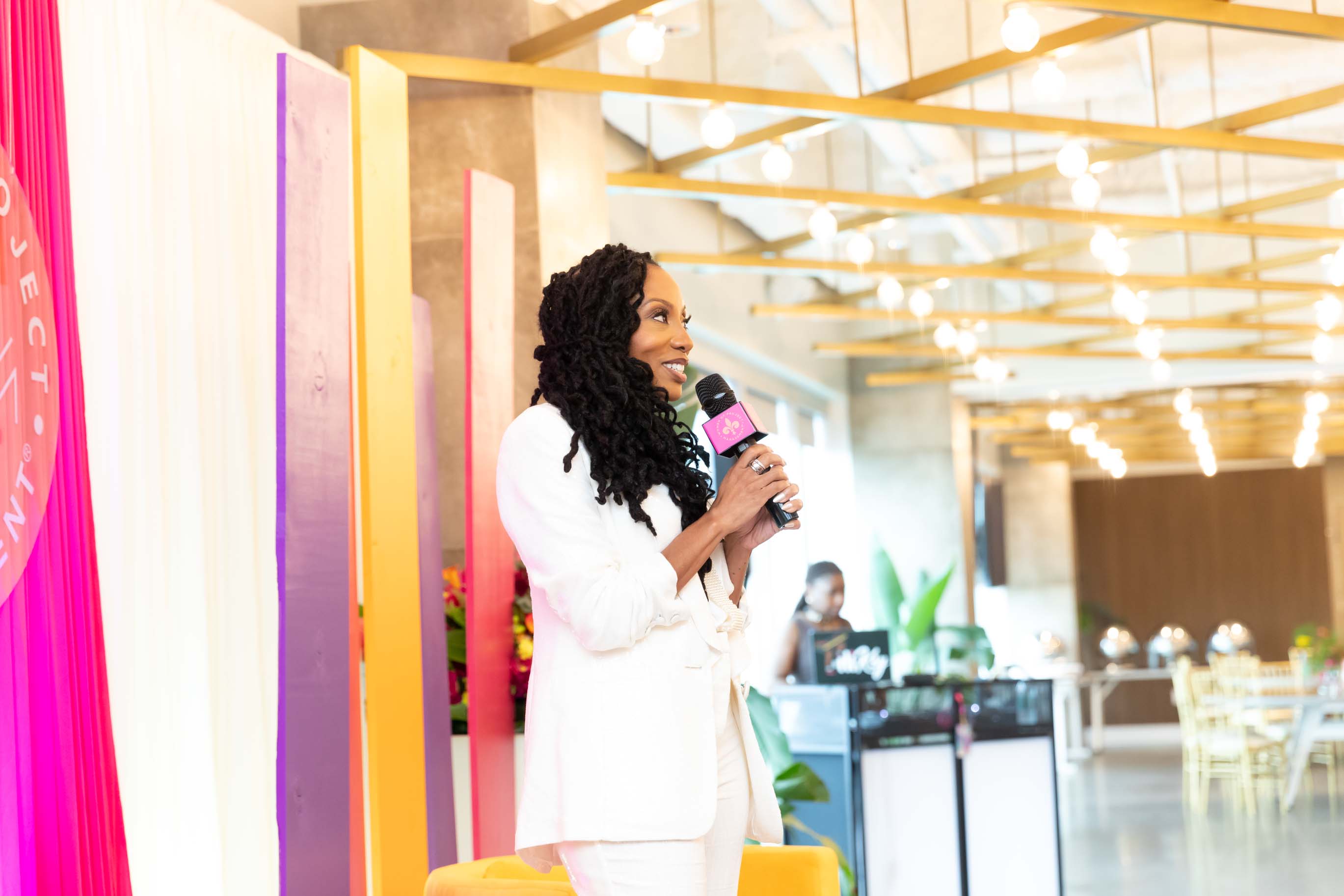Woman with curly hair in white suit holding a microphone, standing indoors with bright lighting and modern decor.