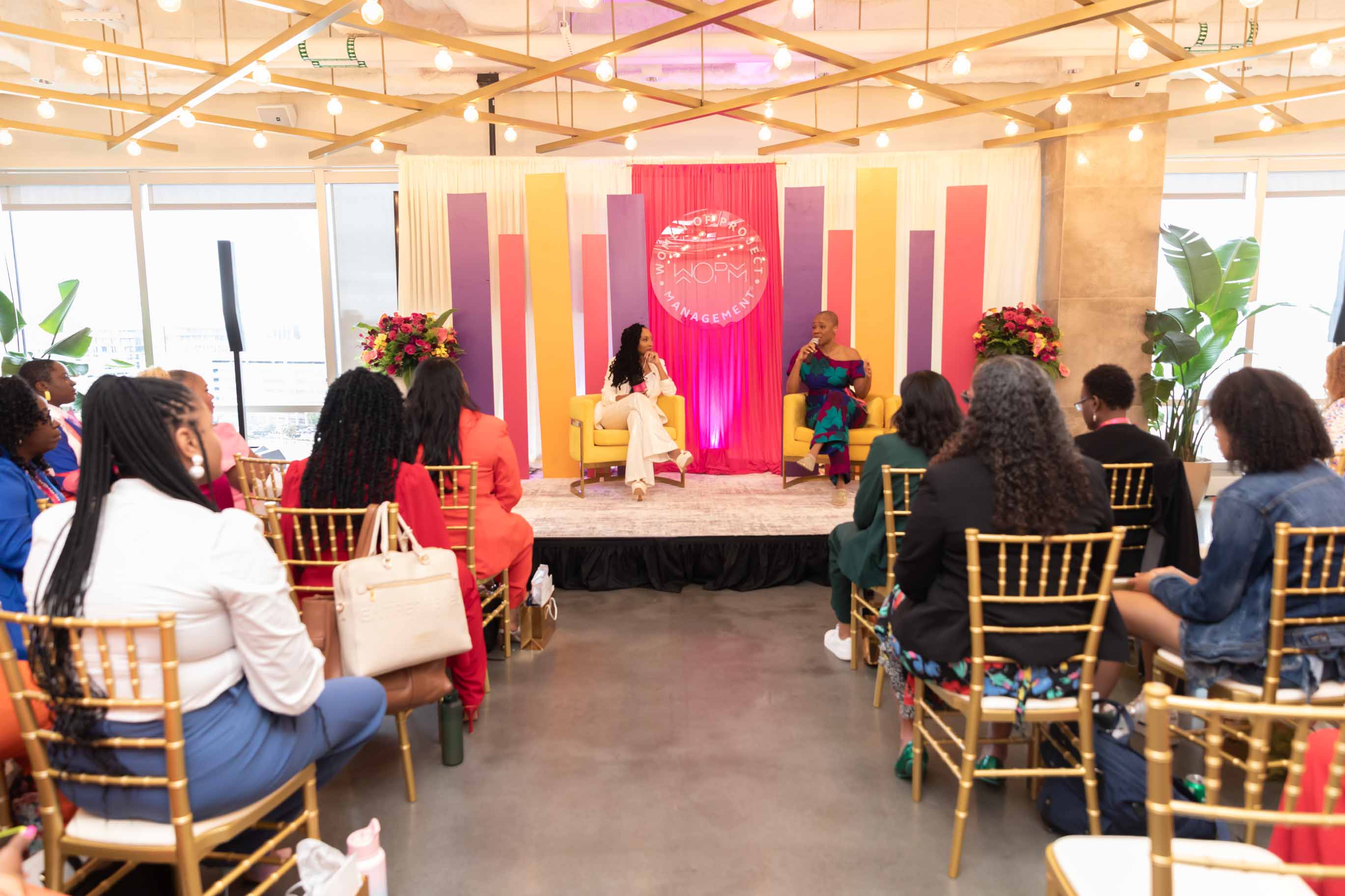 Two women sit on stage in front of an audience in a brightly lit room with colorful backdrop and floral arrangements.