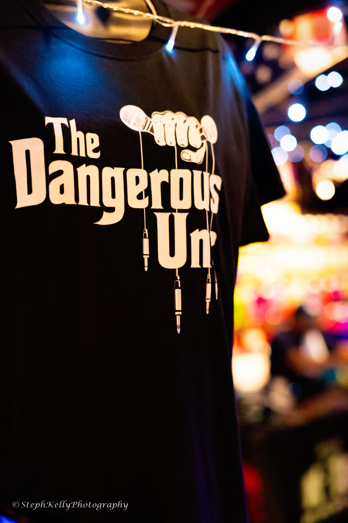 Black T-shirt with white text reading 'The Dangerous Un' hanging in a colorful, blurred background.