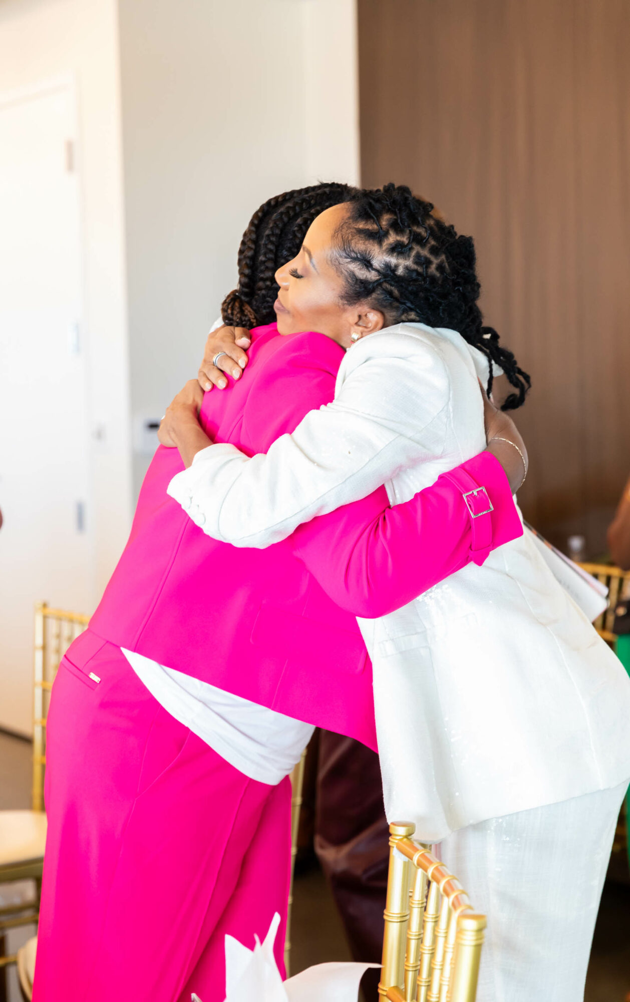 Two women hugging, one in bright pink outfit, the other in white, in an indoor setting with gold chairs.
