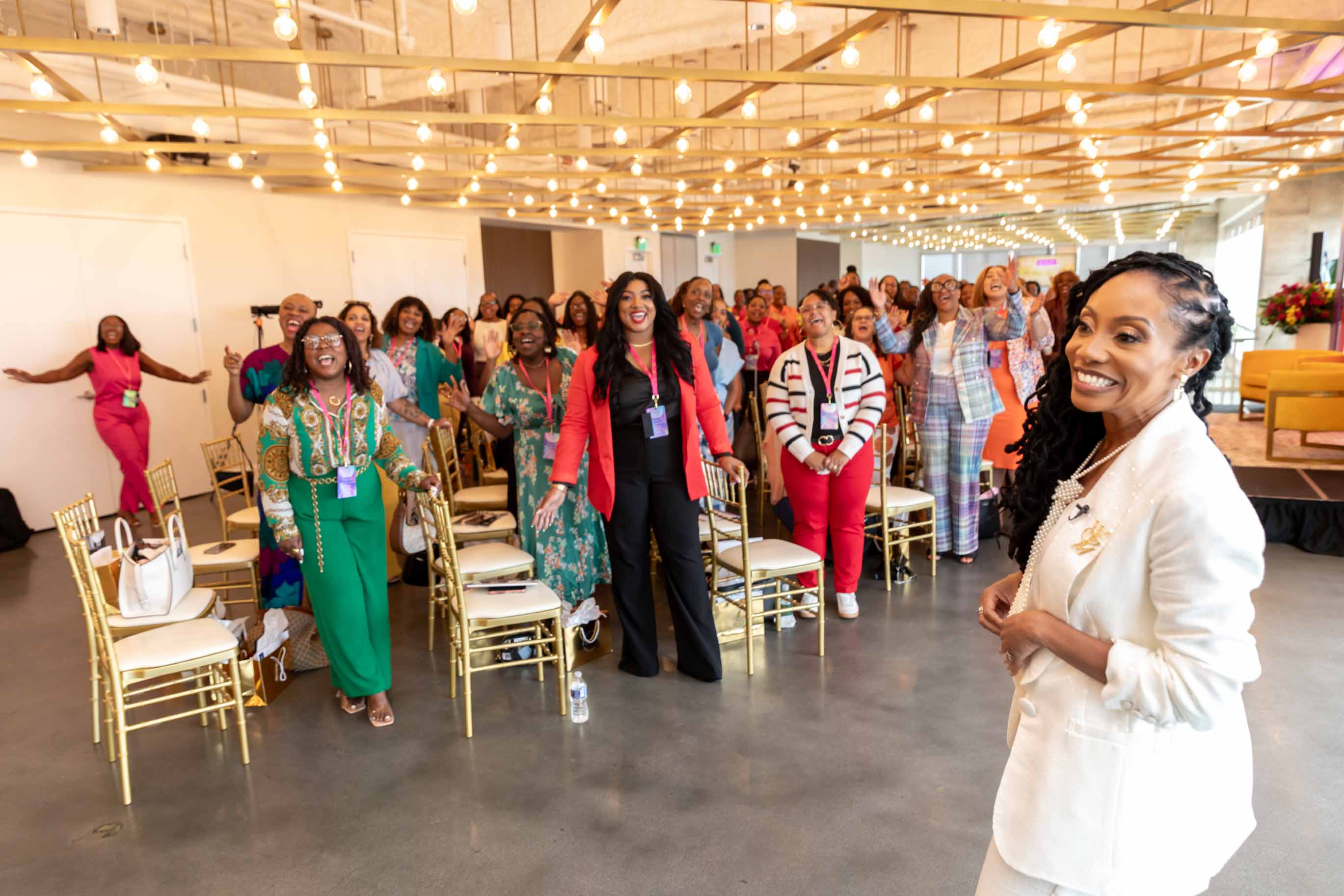 A group of women standing and sitting in a decorated indoor space, smiling and engaging with each other, with a woman in white in the foreground.