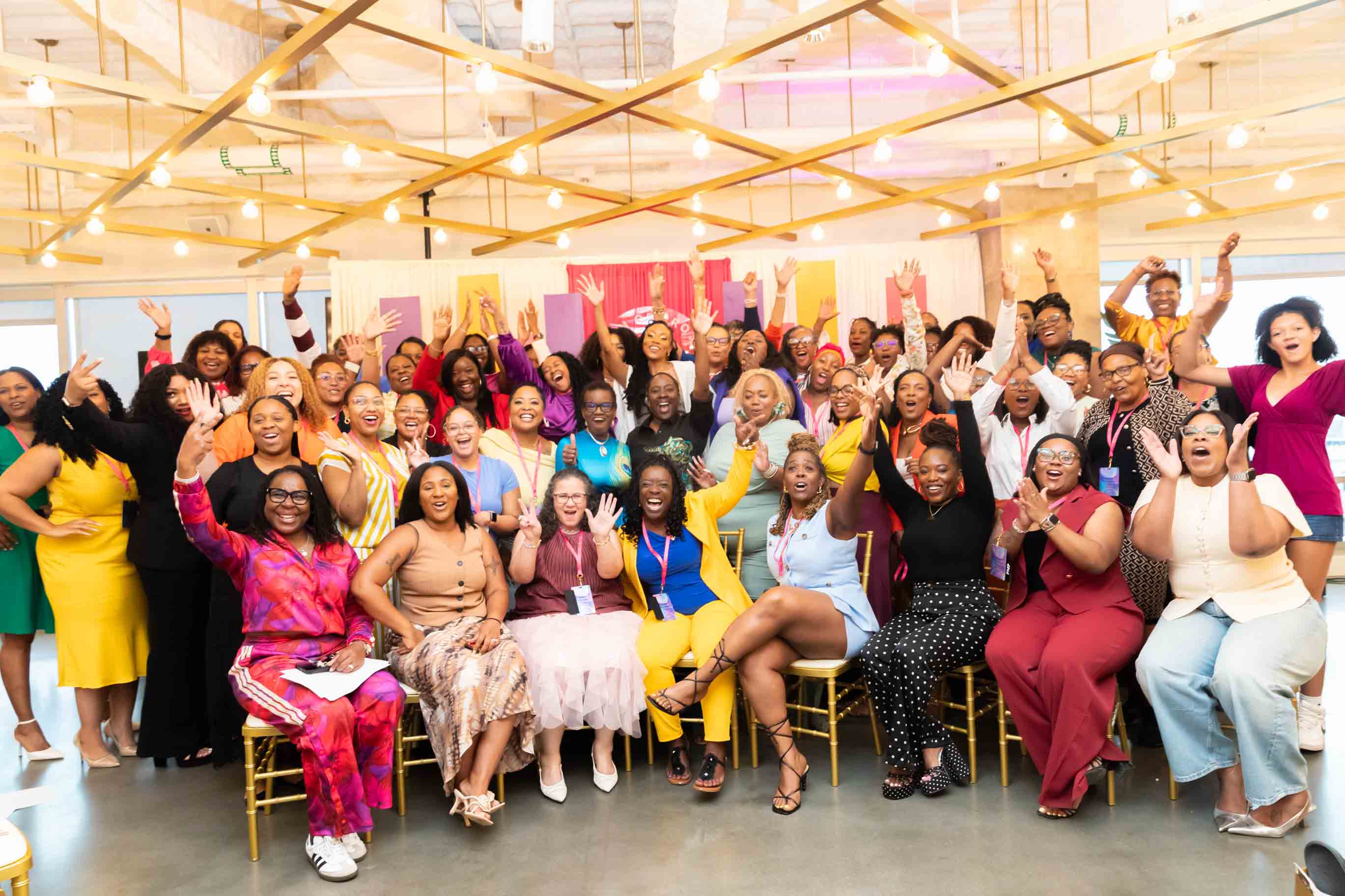 Group of diverse women and men smiling and raising hands in a decorated indoor event space.