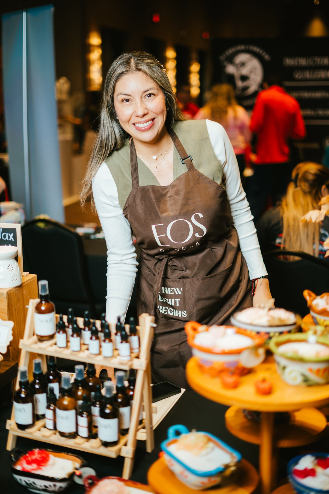 Smiling woman wearing an apron stands behind a table with skincare products and bowls at an indoor event.