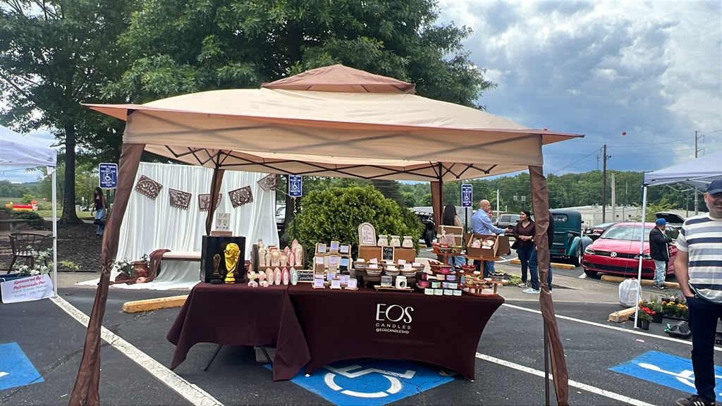 Outdoor market stall with a canopy, displaying various products on a table, with cars and people in the background.