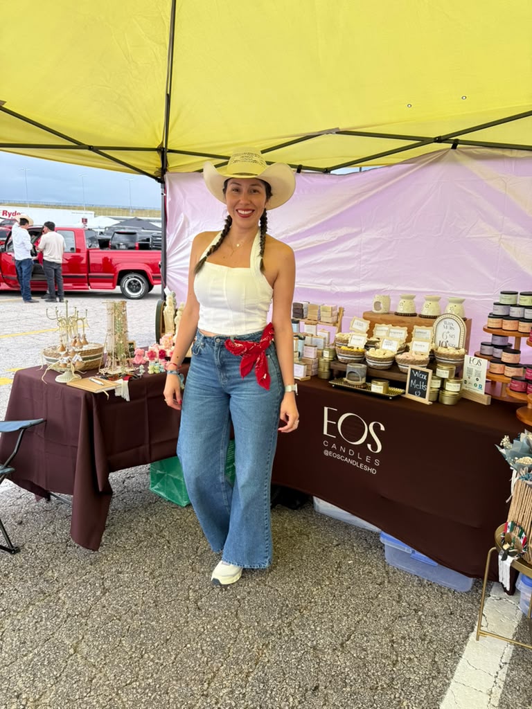 Woman in cowboy hat and casual outfit standing at outdoor market stall with jewelry and accessories, yellow tent overhead.