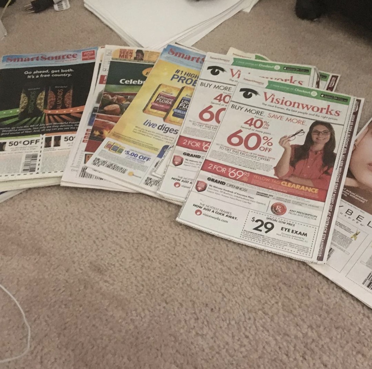 Stack of newspapers and flyers spread on a beige carpet, some overlapping each other, with text and images visible.
