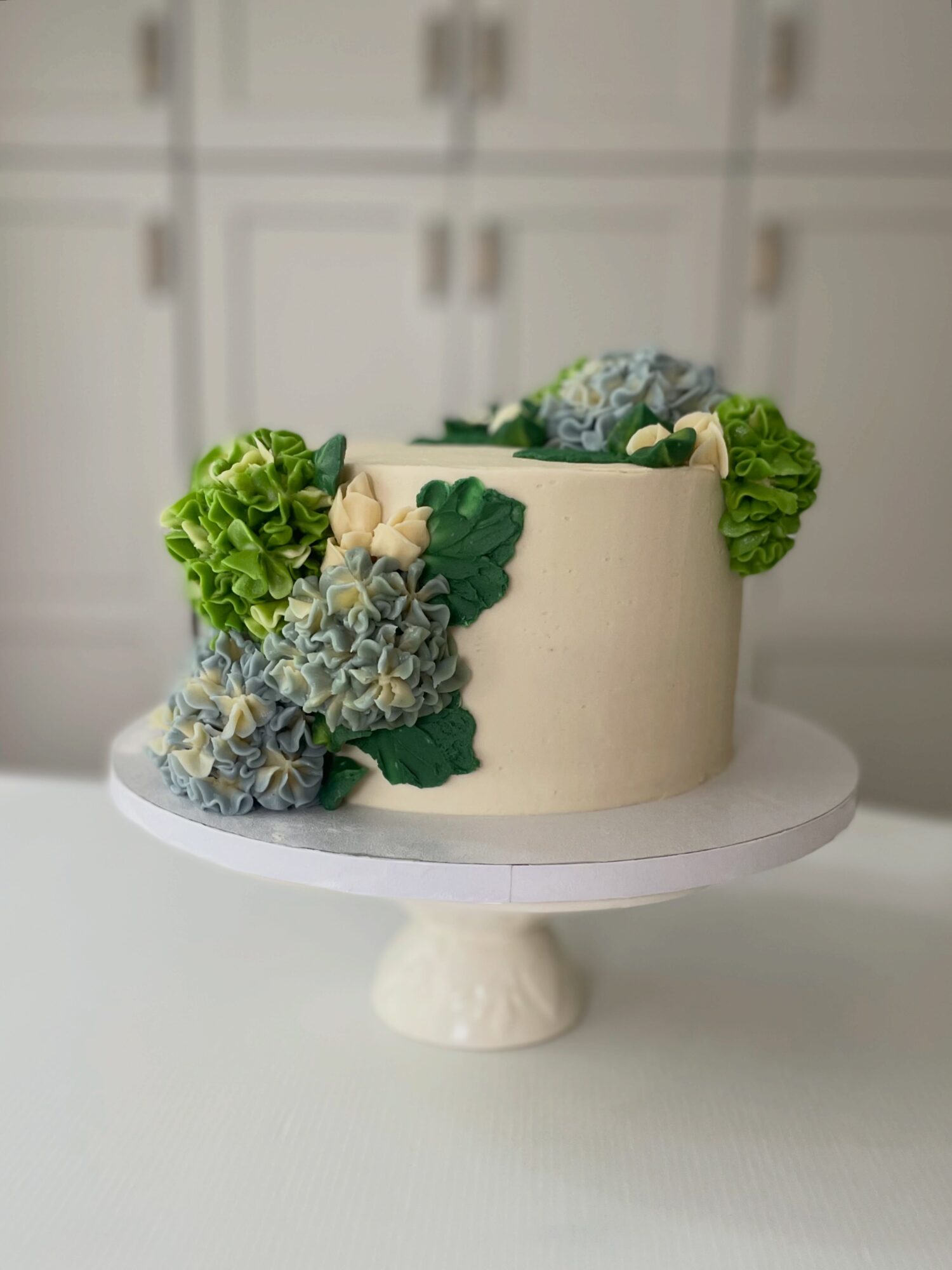 Cream-colored cake decorated with green and blue hydrangea flowers on a white cake stand.
