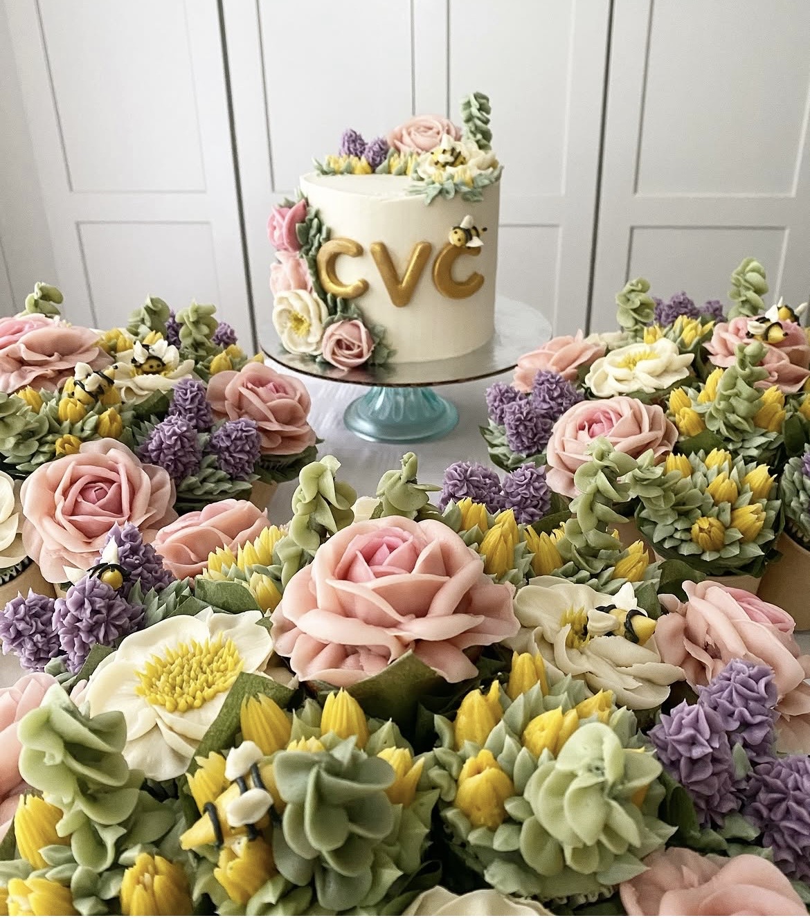 A white cake with pink, purple, and yellow flowers on top, surrounded by floral arrangements with roses, daisies, and other flowers, on a table.