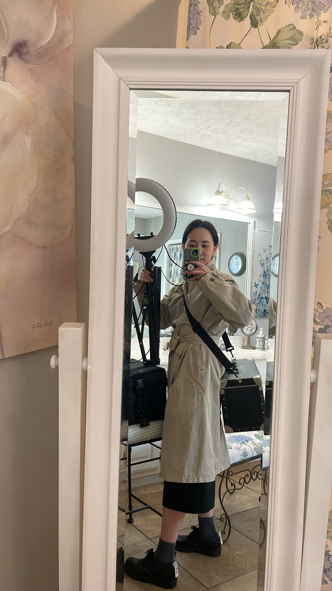 Person taking a mirror selfie in a room with floral wallpaper, wearing a beige coat, black pants, and boots.