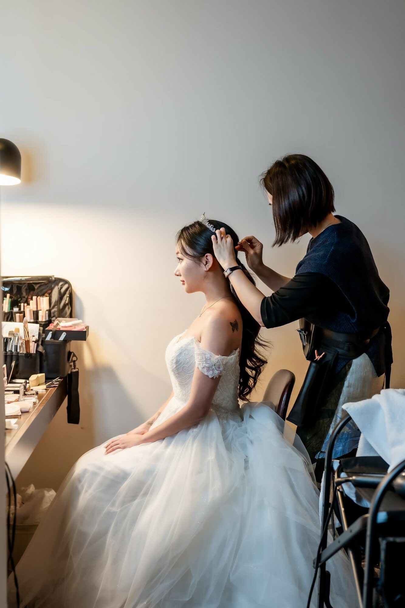 Woman in wedding dress getting her hair styled by a stylist in a room with makeup tools and a mirror.