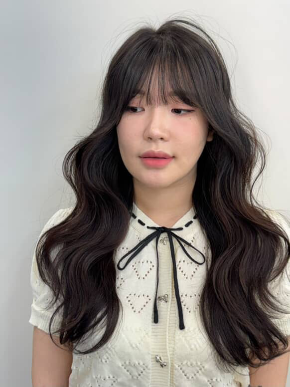 Young woman with long, wavy dark hair and bangs, wearing a white blouse with a black ribbon, looking to the side.