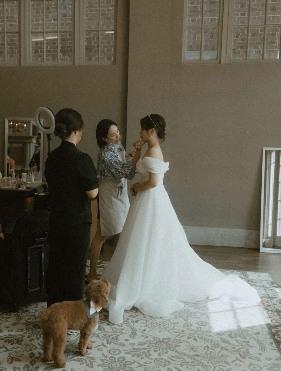 Three women and a dog in a room with large windows, one woman in a wedding dress, two women in dark clothing, dog on a leash.