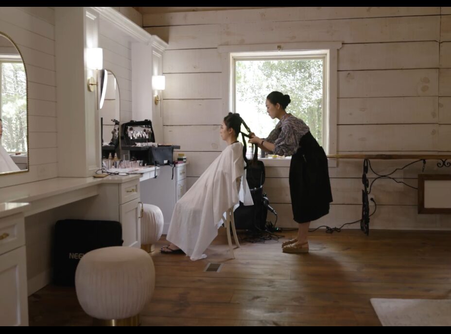 Person getting hair styled by another person in a room with white wooden walls and a window, with a stool and mirror nearby.
