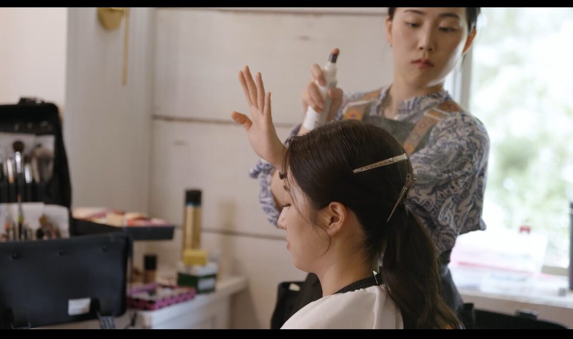 A woman with long dark hair is getting her hair styled by a stylist in a room with natural light.