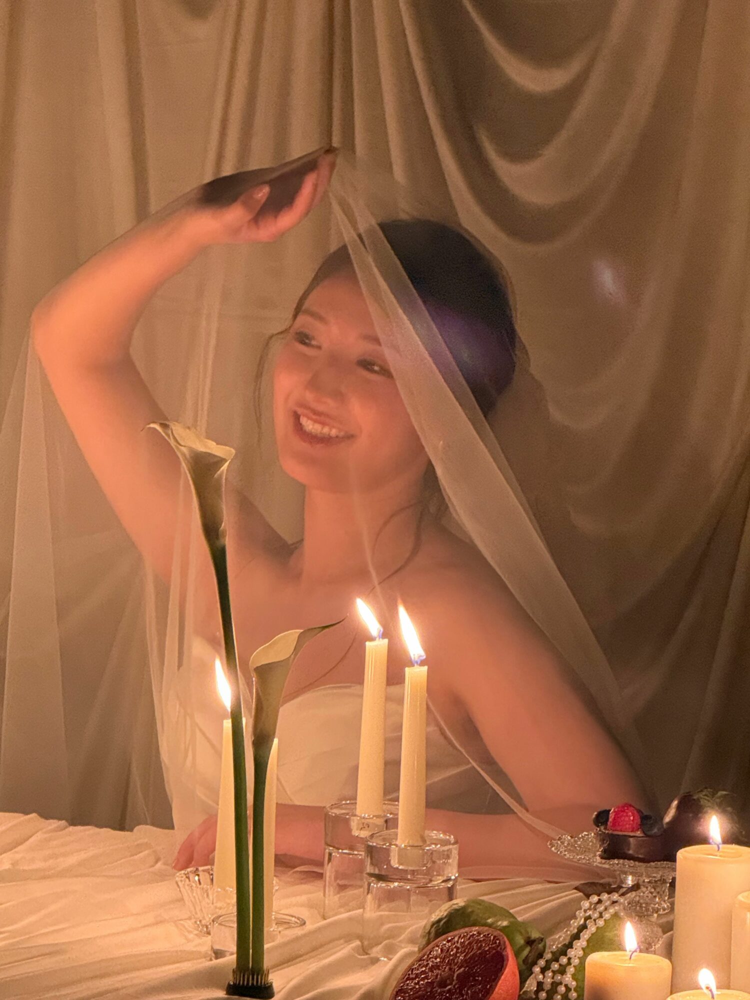 Woman smiling behind a sheer curtain, holding a phone, with a table of candles and flowers in front.