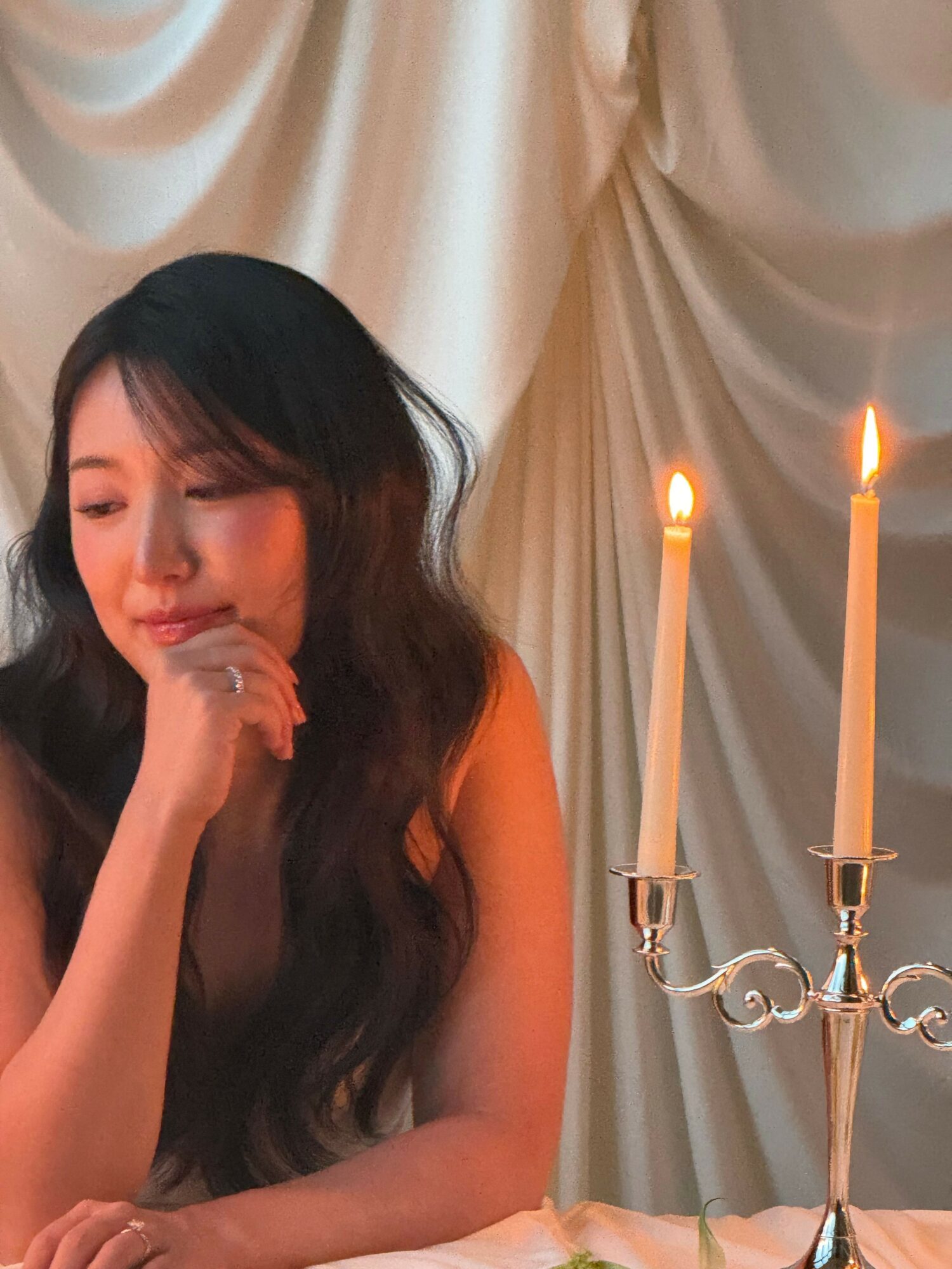 Woman with long dark hair resting her chin on her hand, smiling softly, in front of a curtain and chandelier.