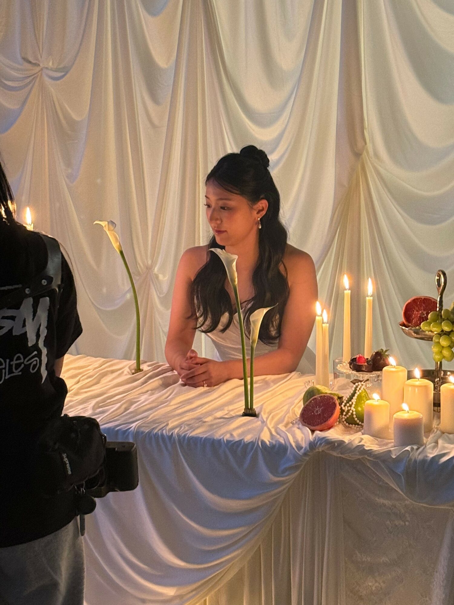 Woman with long dark hair leaning on a table decorated with candles and flowers, with a draped backdrop behind her.