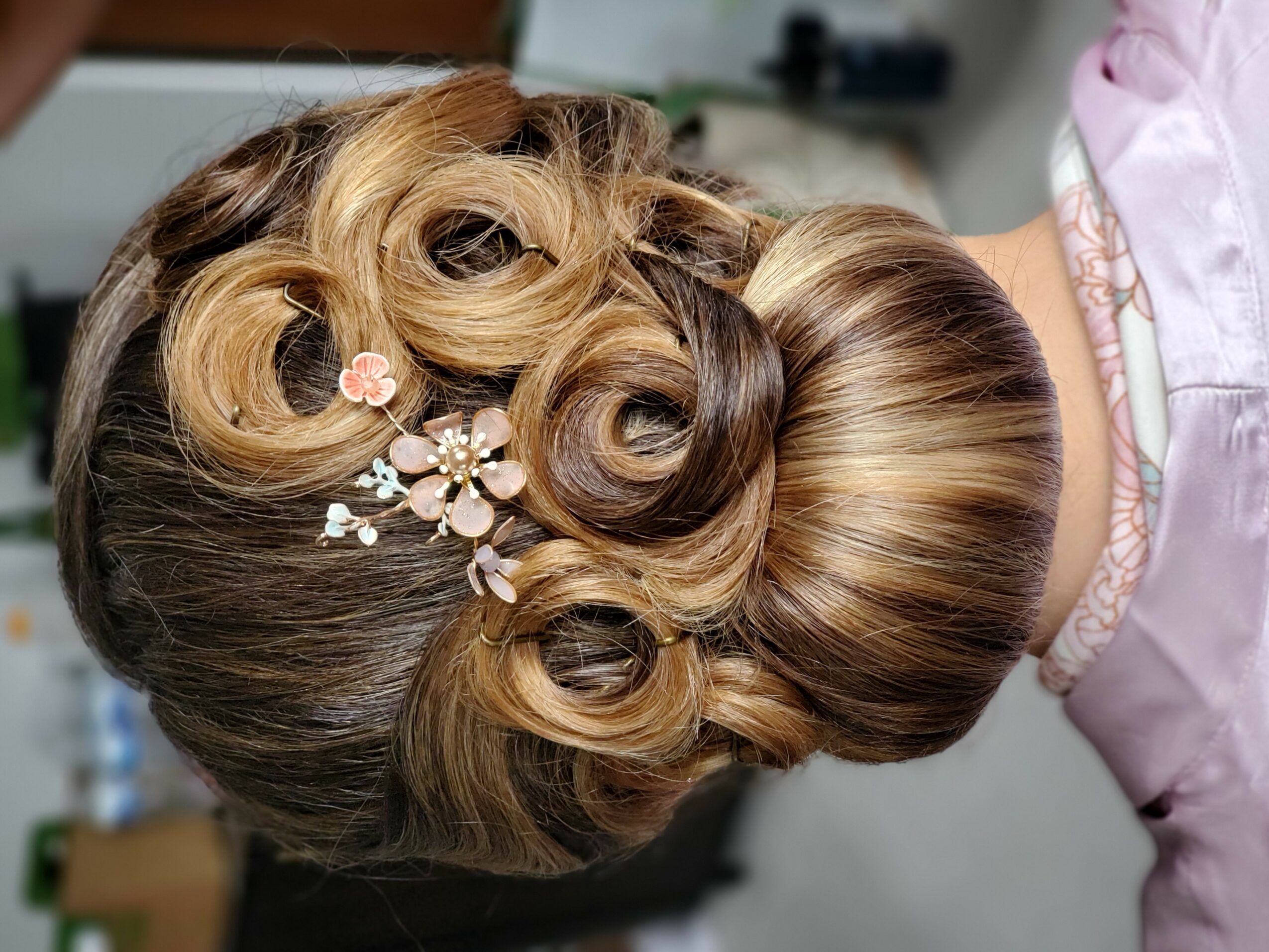 Intricate updo hairstyle with curls and floral hair accessories, viewed from the back, with a person wearing a pink garment.