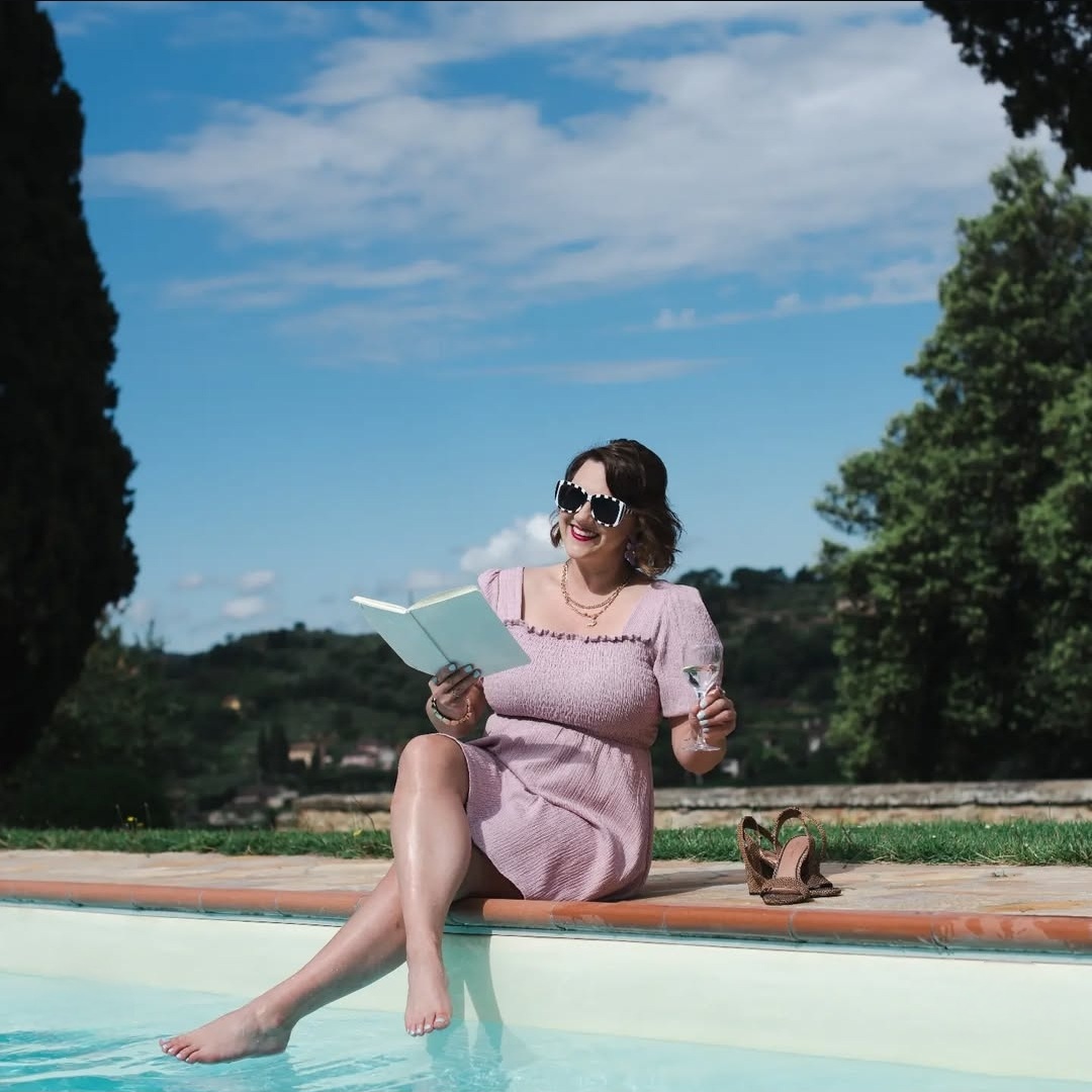 Woman in pink dress sitting on pool edge reading a book outdoors, with trees and blue sky in background.