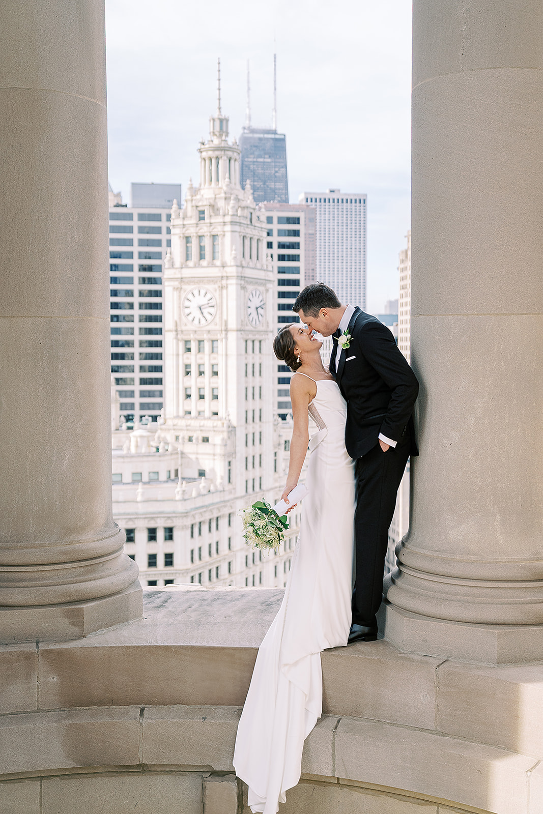 Bride and groom sharing a kiss between large columns with city skyscrapers in the background.