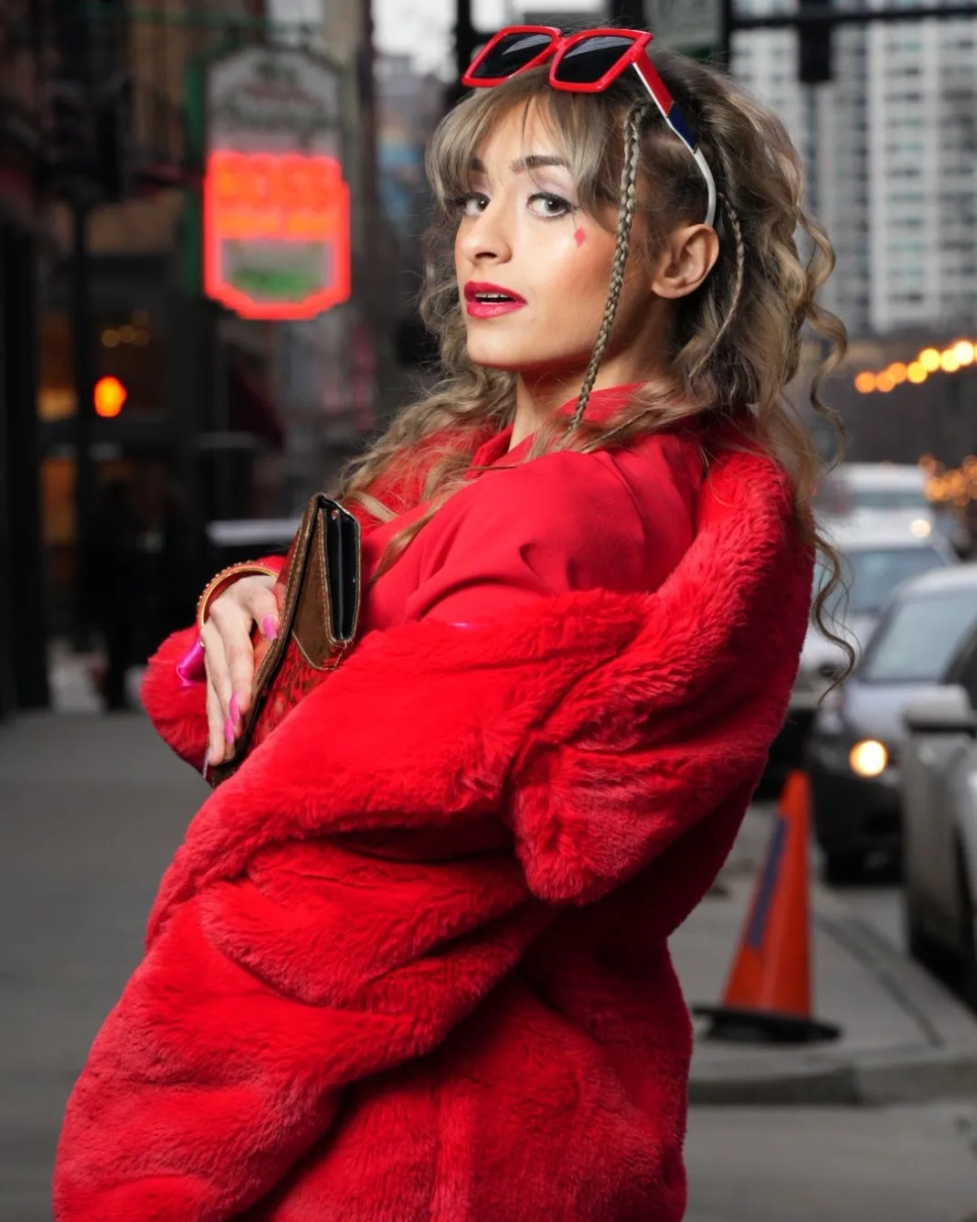 Young woman with wavy hair in a red coat and sunglasses on her head, standing on city street with cars and buildings.