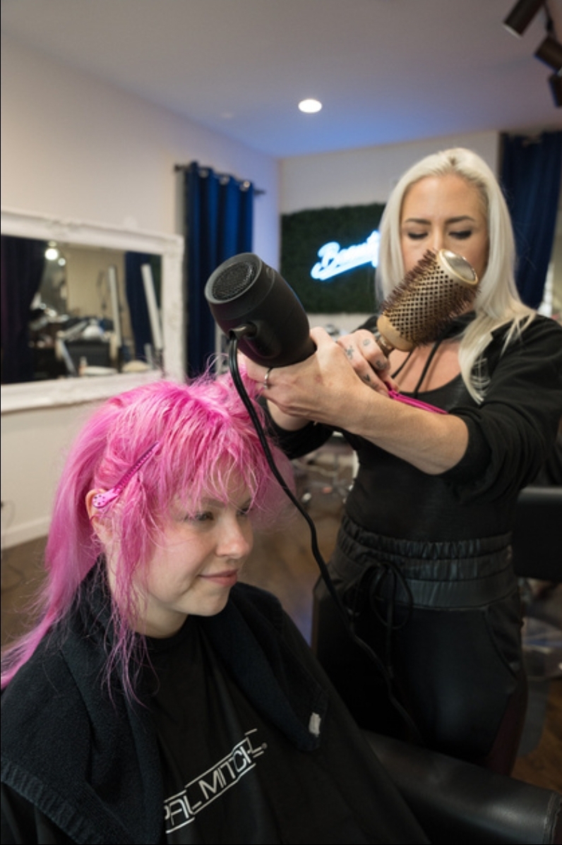 Hairdresser with blonde hair and face mask blow-drying a person's pink hair in a salon.