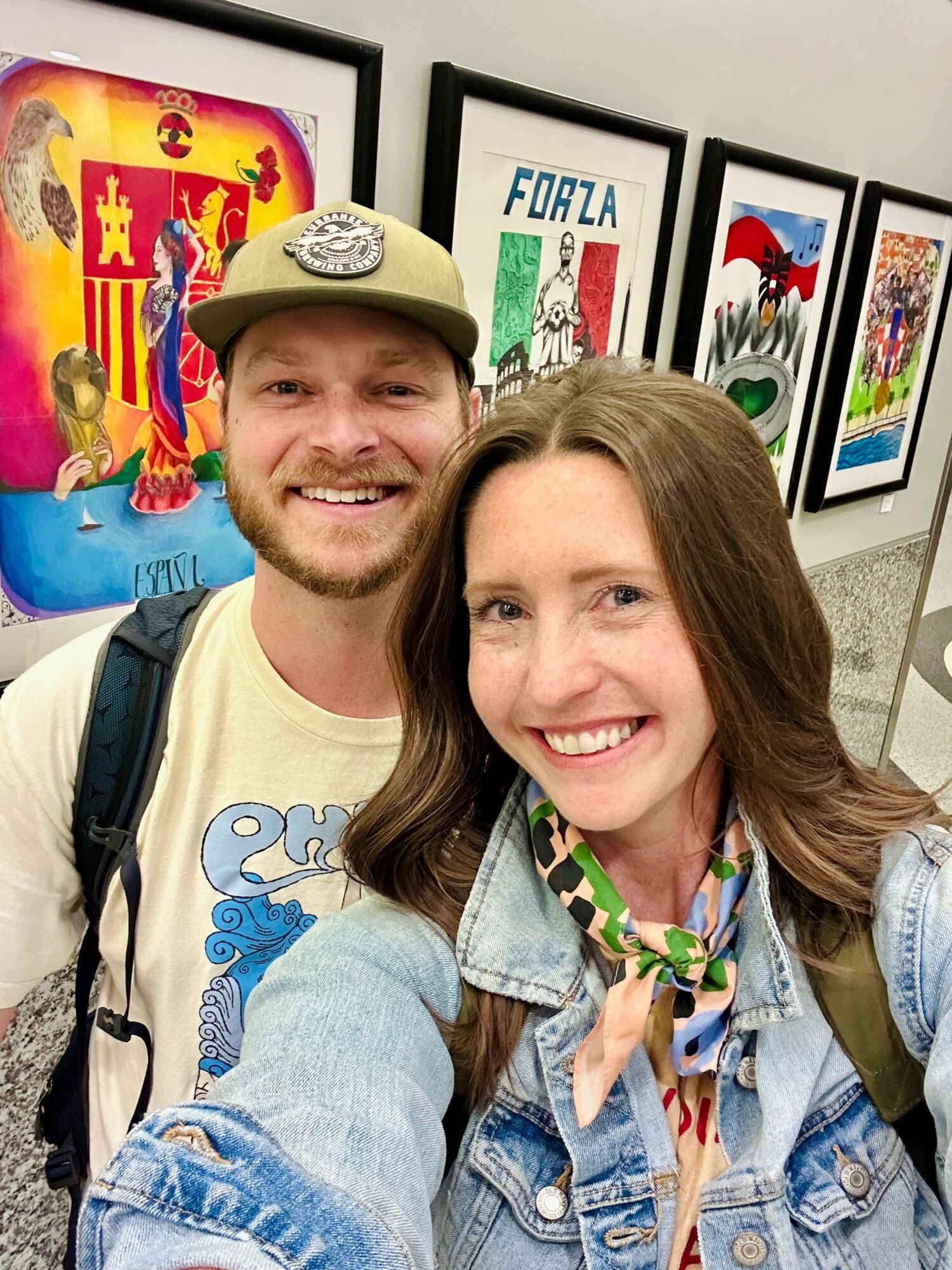 Two smiling people taking a selfie in front of colorful framed artwork on a wall.