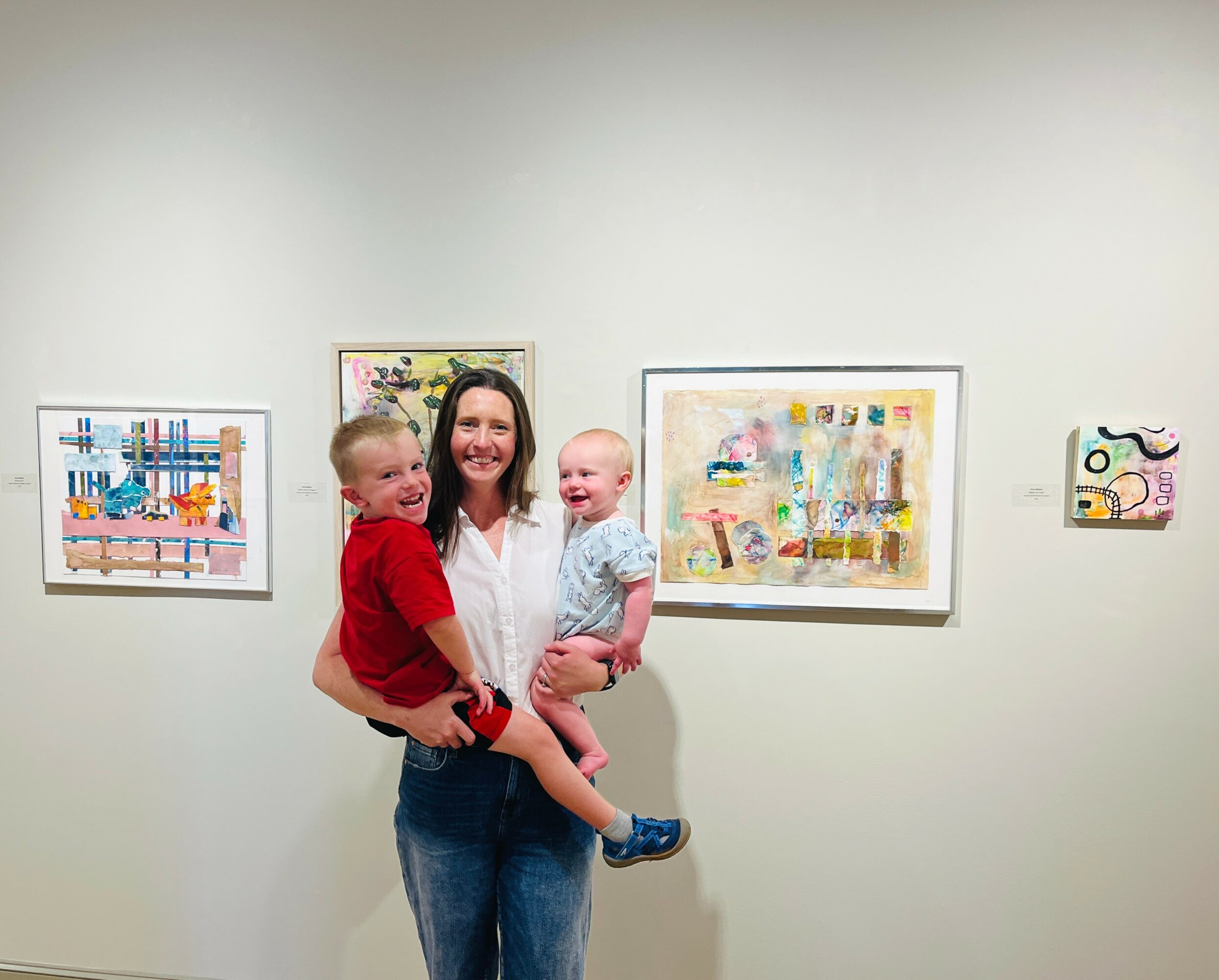 Woman holding two children in front of colorful artwork on gallery wall.