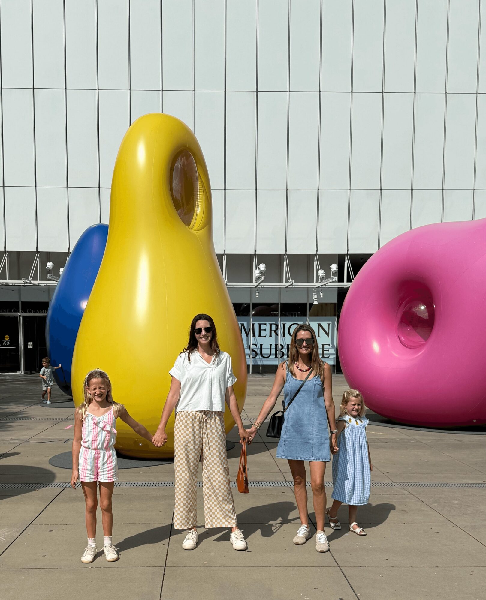 Four people, two adults and two children, holding hands in front of large colorful abstract sculptures, on a sidewalk outside a building.