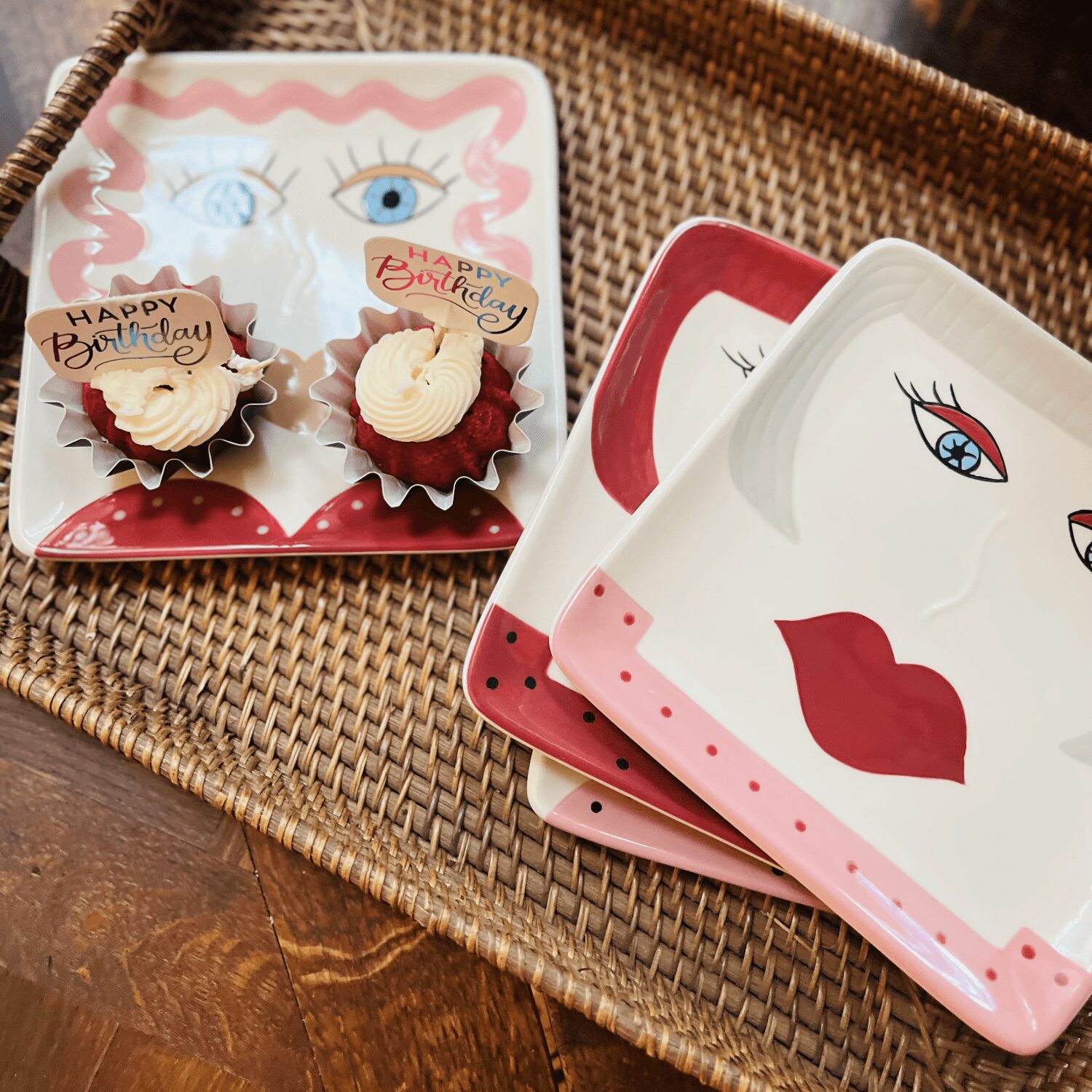 Decorative plates with eye and lip illustrations, cupcakes with birthday toppers, on a woven tray.