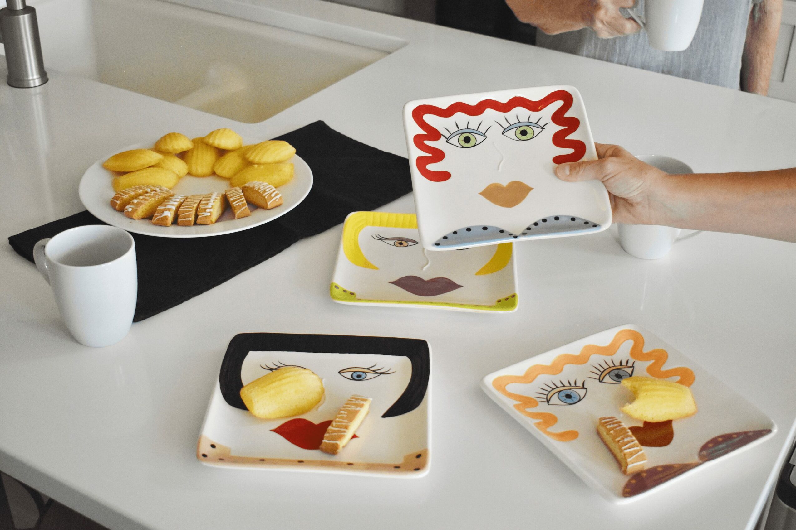 Person holding a plate with a face design, three decorated plates with cookies, a cup, and a black cloth on a white table.