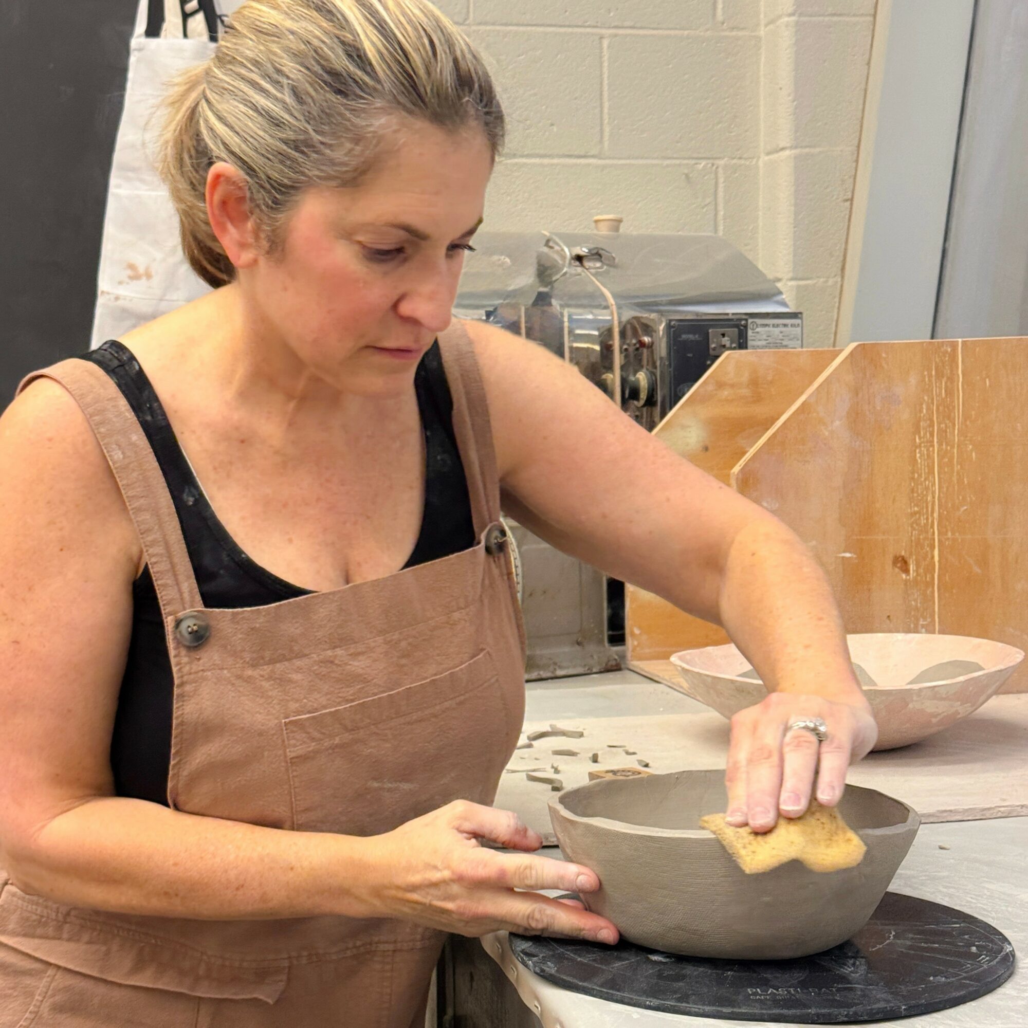 Woman shaping a ceramic bowl in a workshop, wearing an apron, with pottery tools and equipment in the background.