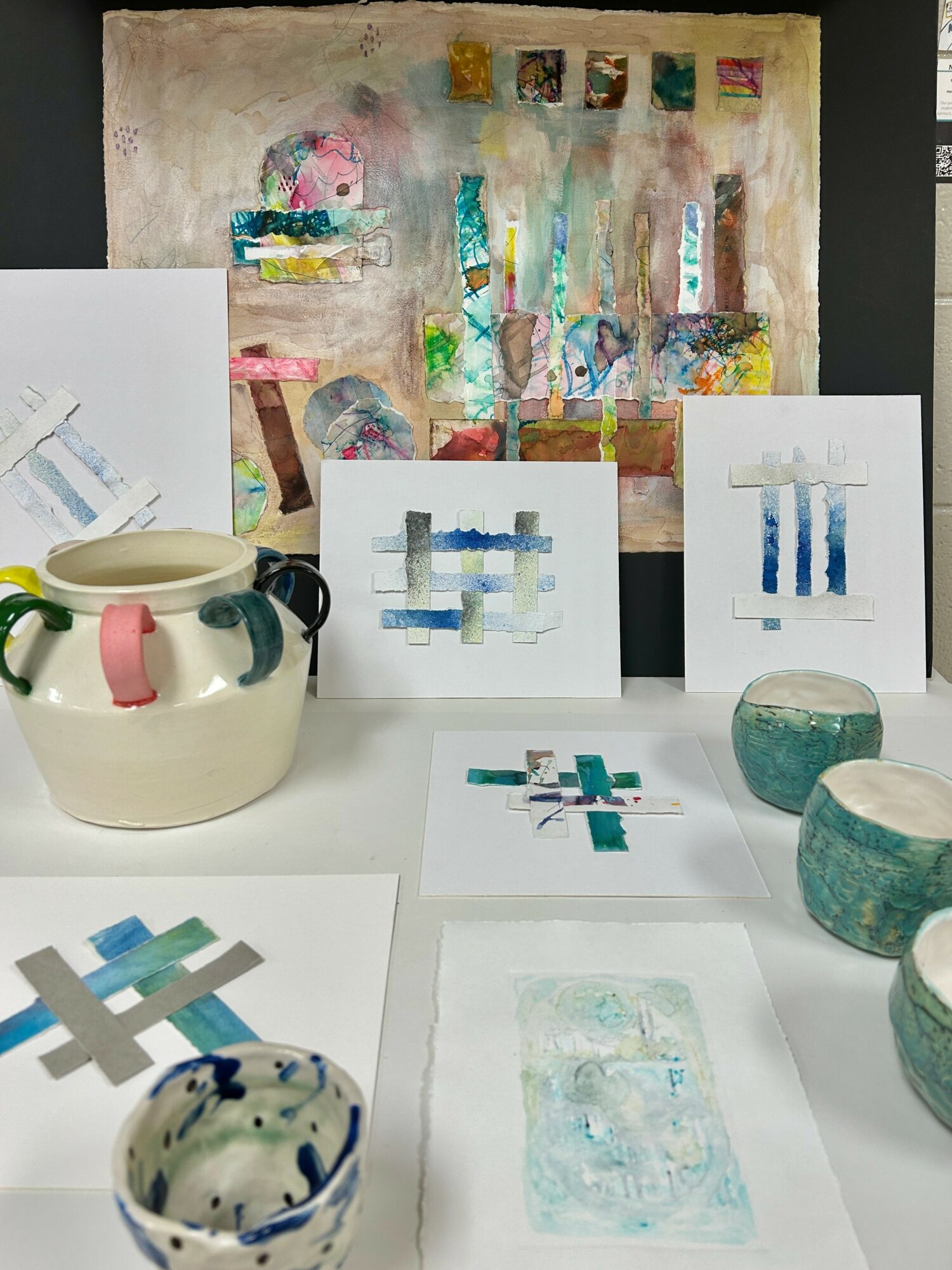 Display of abstract watercolor artworks, ceramic bowls, and small painted objects on a table.
