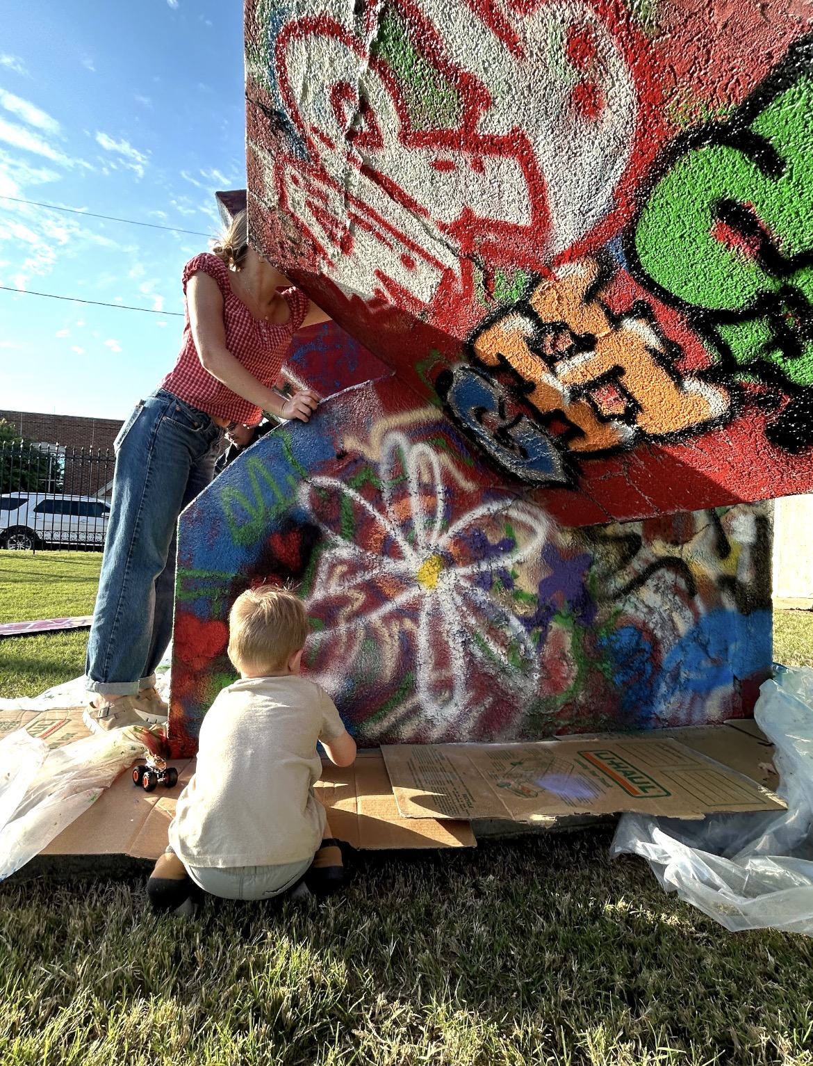 Two children painting graffiti on a wall outdoors, with one standing and the other sitting, under a sunny sky.
