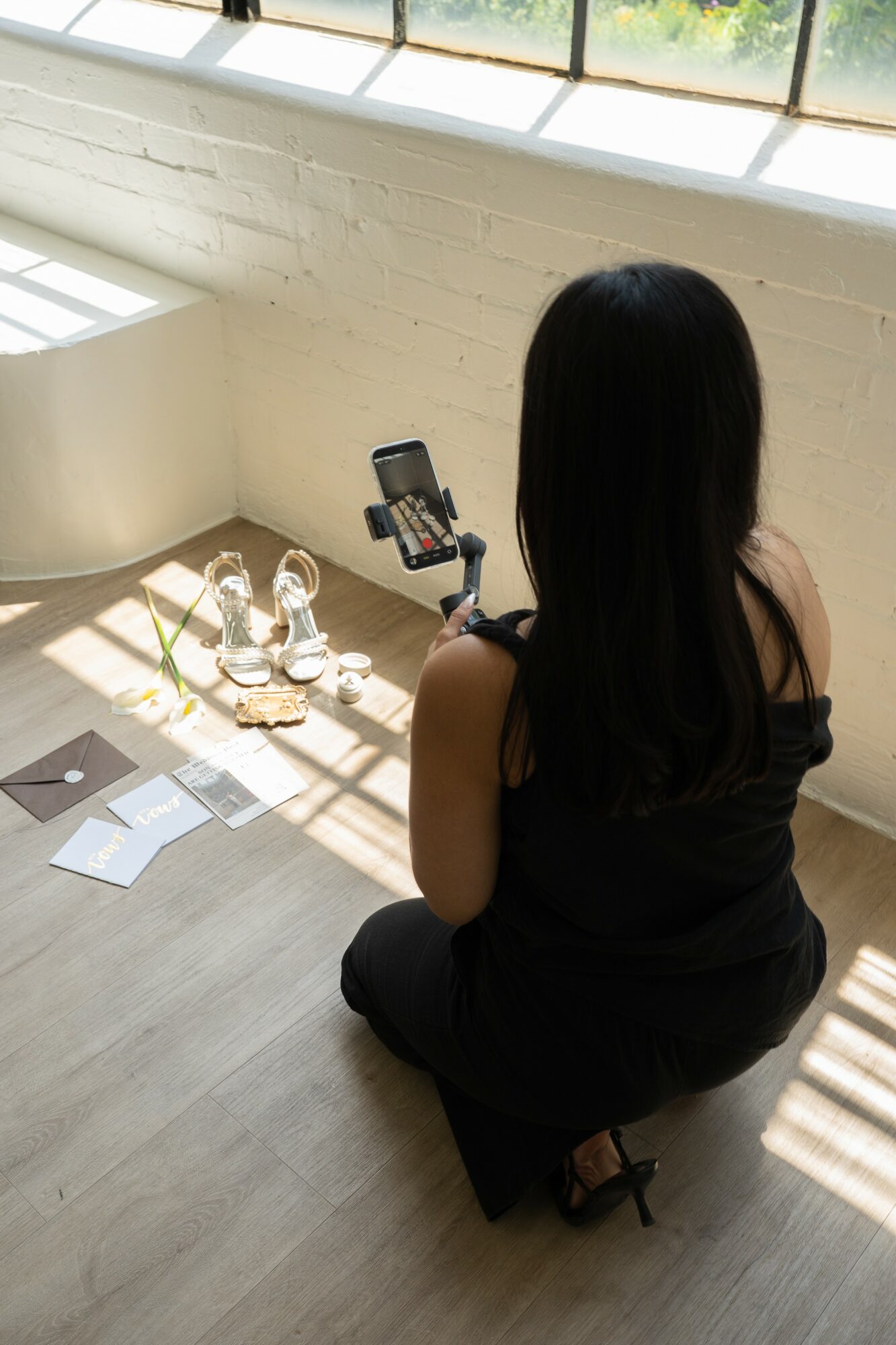 Woman with long dark hair kneeling on floor, looking at camera on tripod, sunlight streaming through window, bottles and papers nearby.