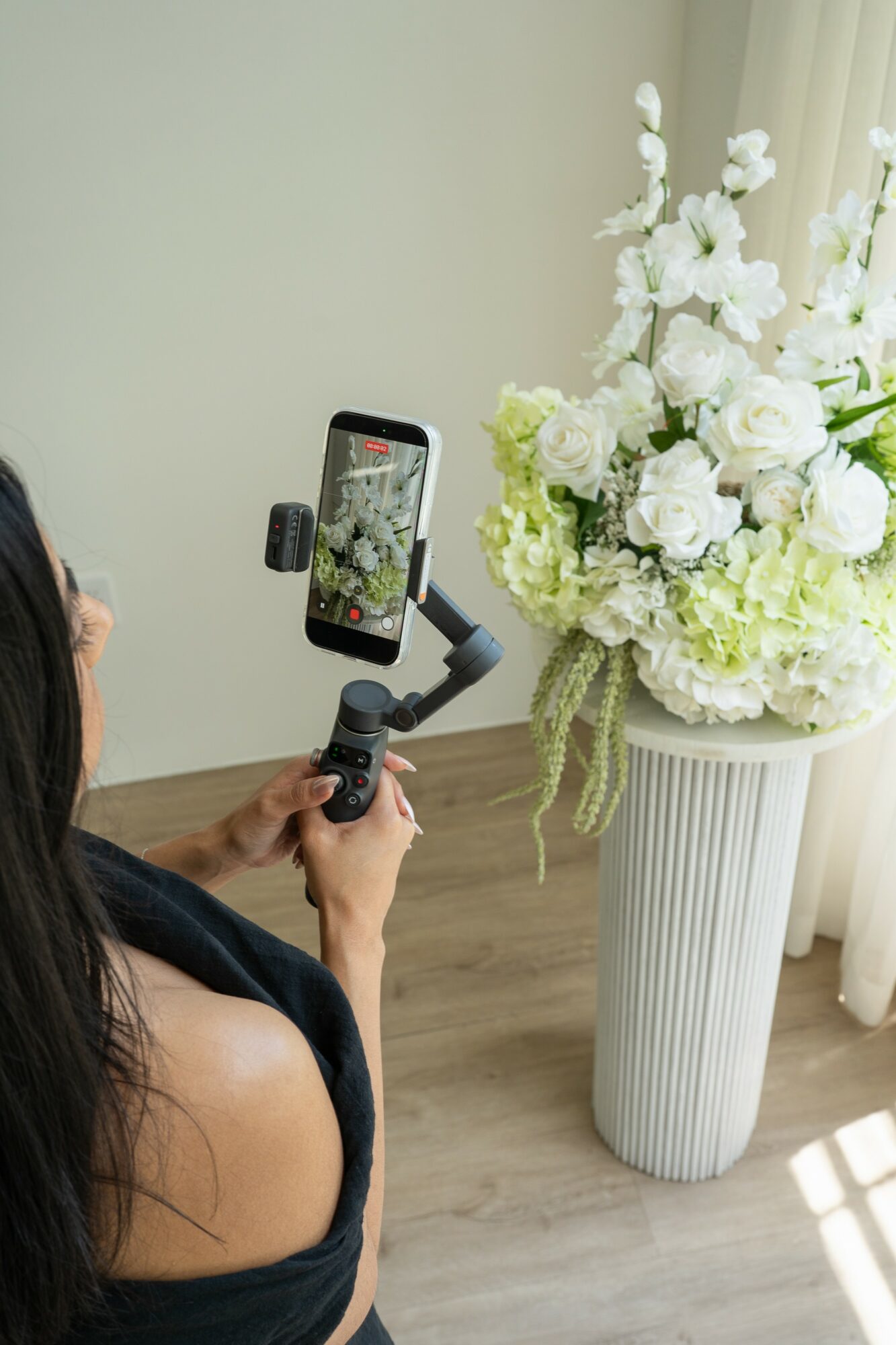 Person holding a camera stabilizer taking a photo of a large white flower arrangement in a tall vase, sunlight on the floor.