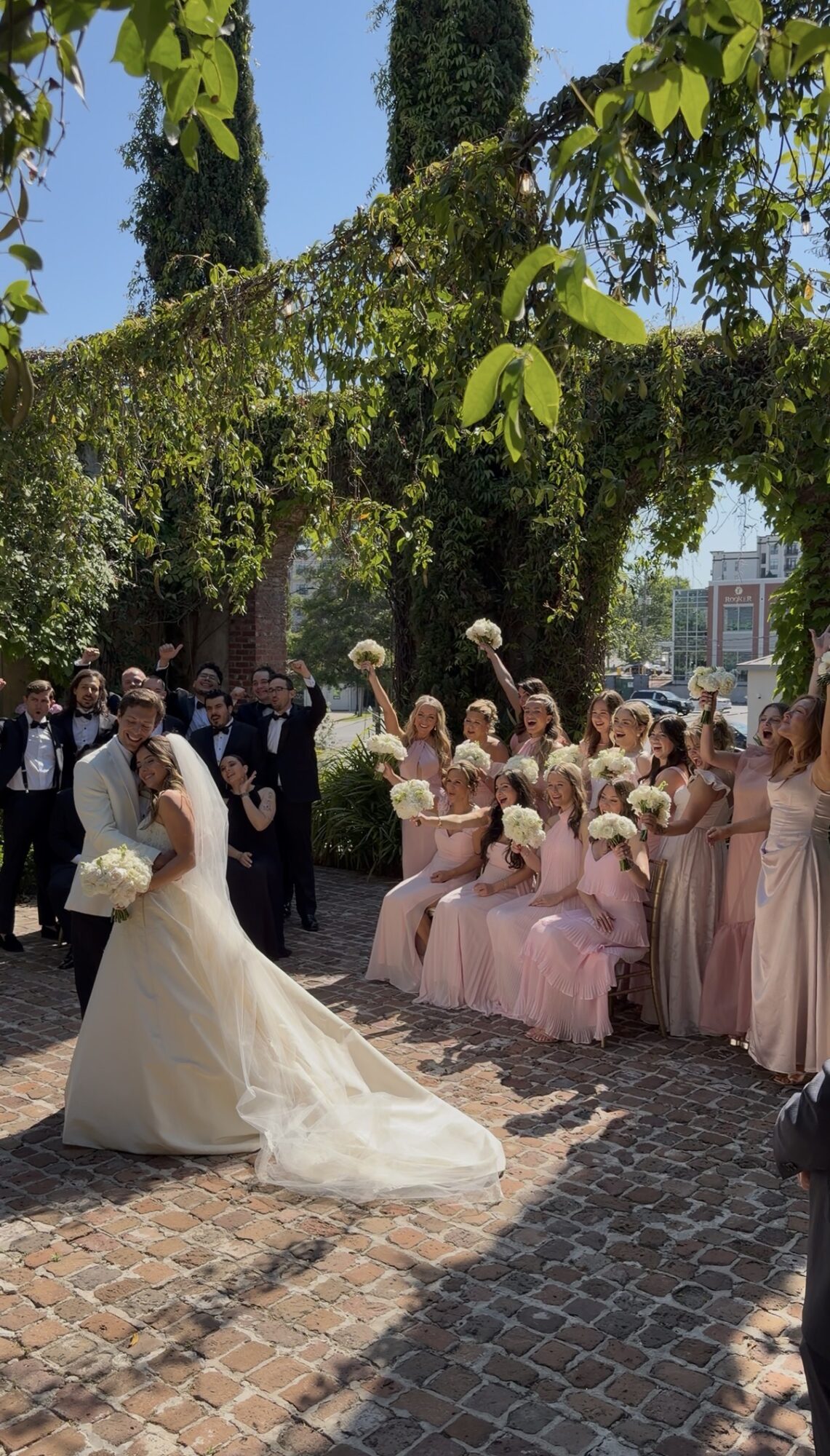 Bride in a white gown with a long train, walking on cobblestone, surrounded by bridesmaids in pink dresses and guests outdoors.