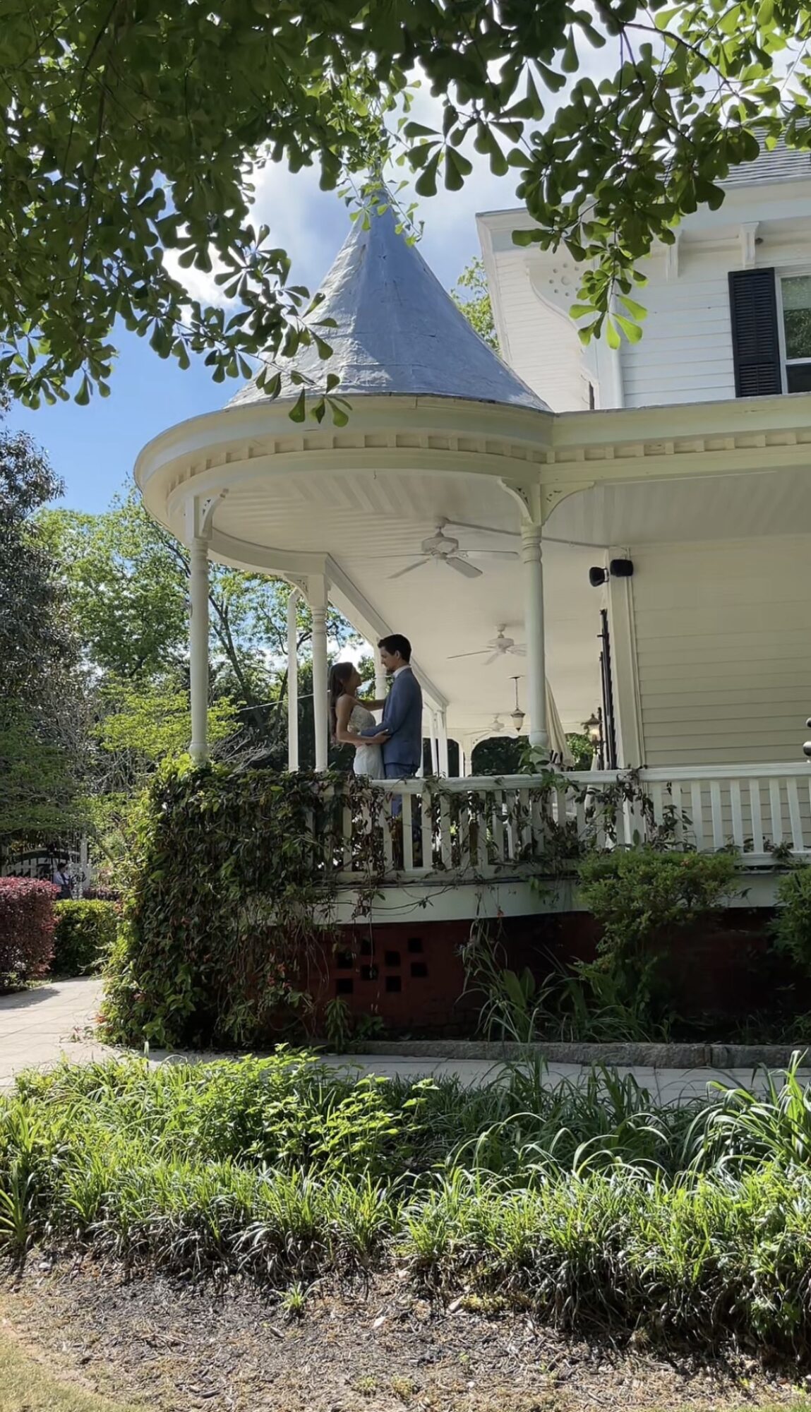 Two people stand on a porch of a white house with a turret, surrounded by greenery and trees.