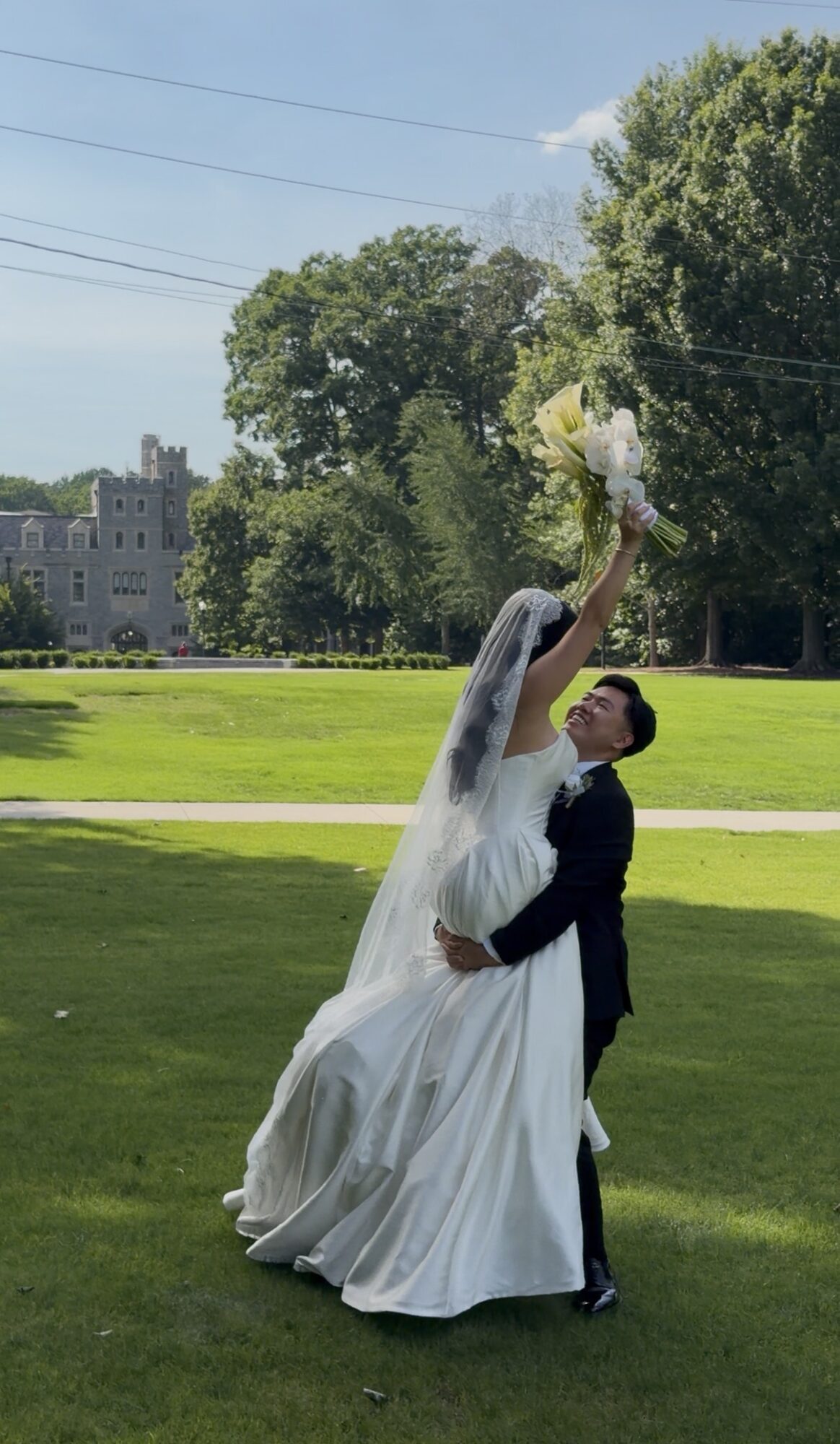 Bride and groom dancing outdoors on grass, bride holding bouquet overhead, trees and a building in background.
