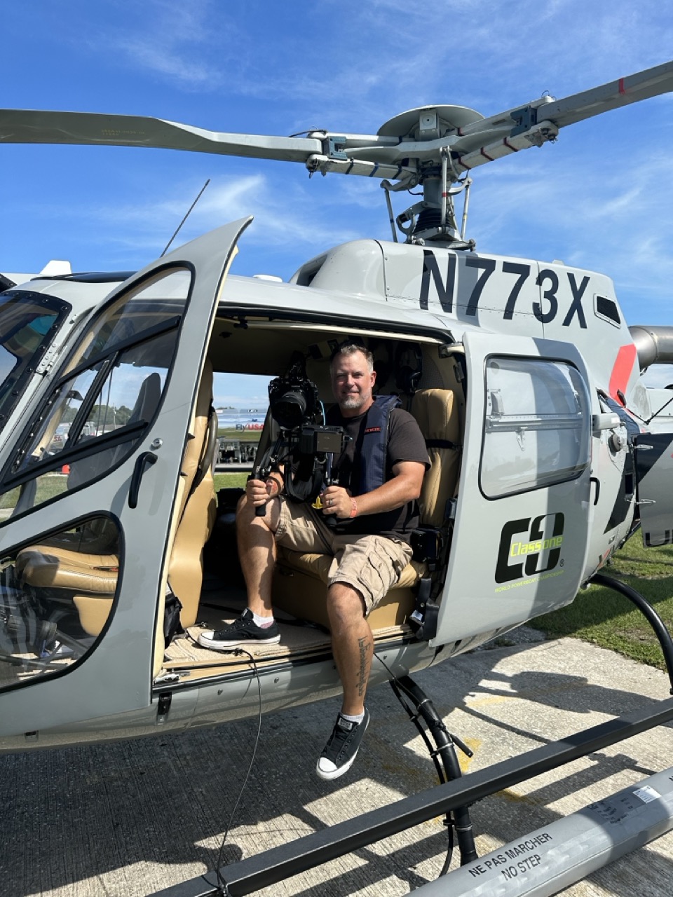 Person sitting inside a helicopter with open door, holding a camera, on a sunny day with blue sky.