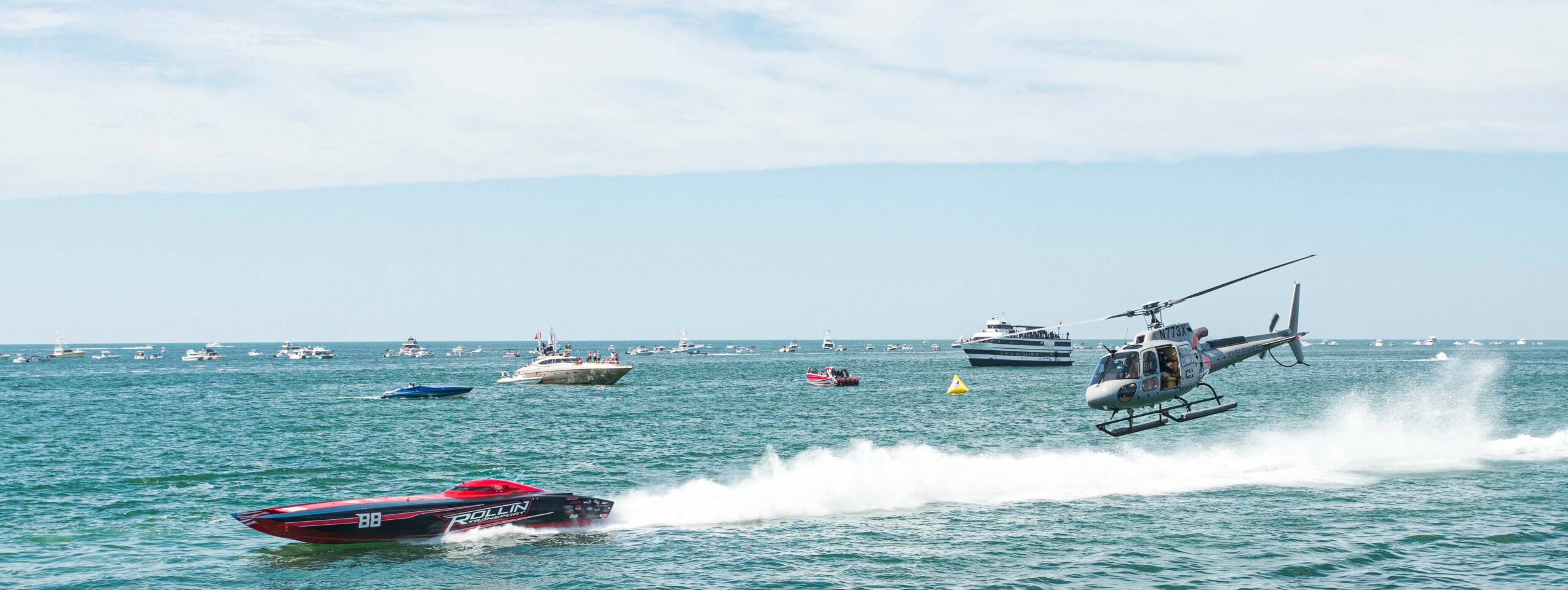 Helicopter flying over water with boats and a speedboat in the foreground, waves and sky visible.