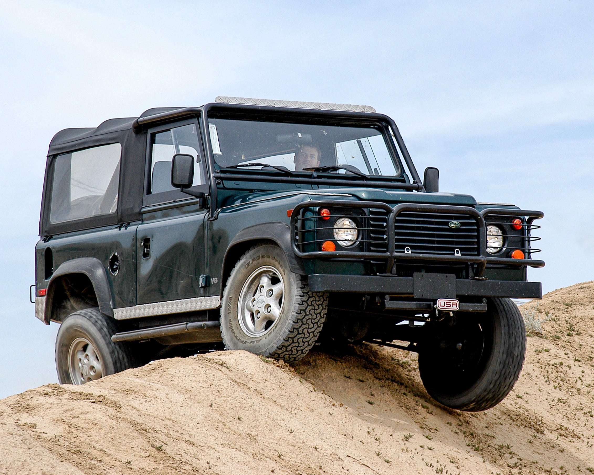 Dark green off-road vehicle climbing sandy terrain with a black soft top and rugged tires.