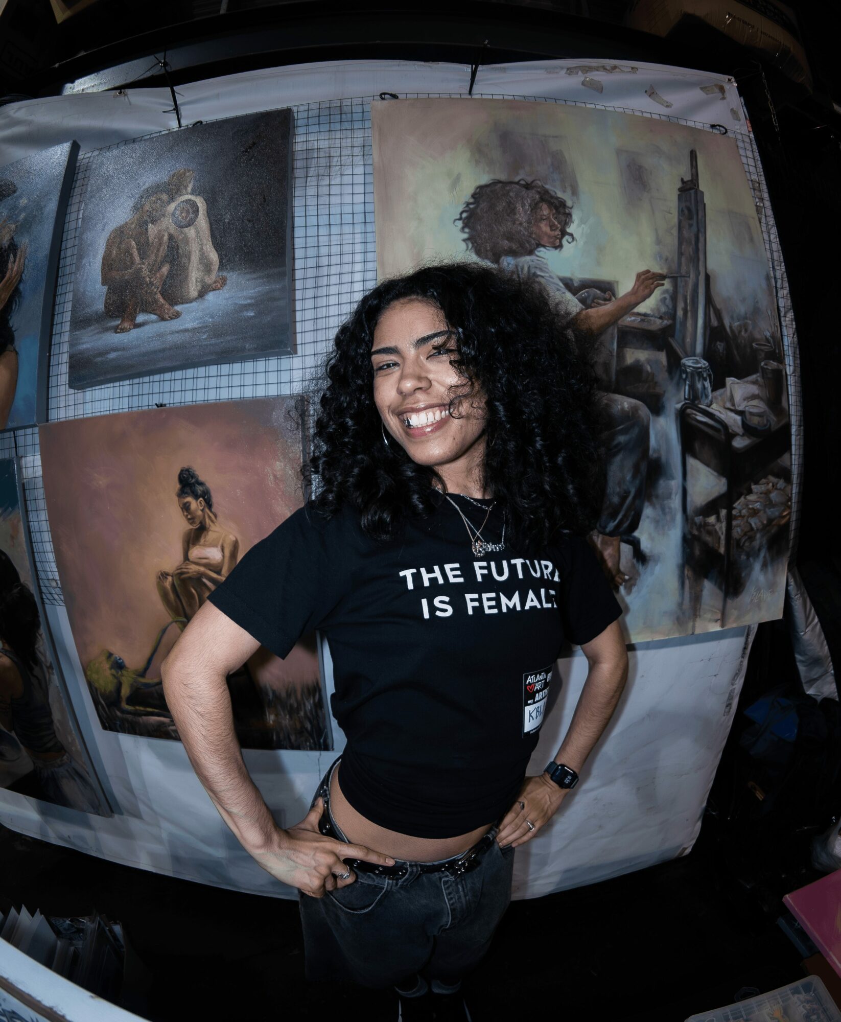 Young woman with curly hair smiling, wearing a black T-shirt that says 'The future is female,' standing in front of paintings.