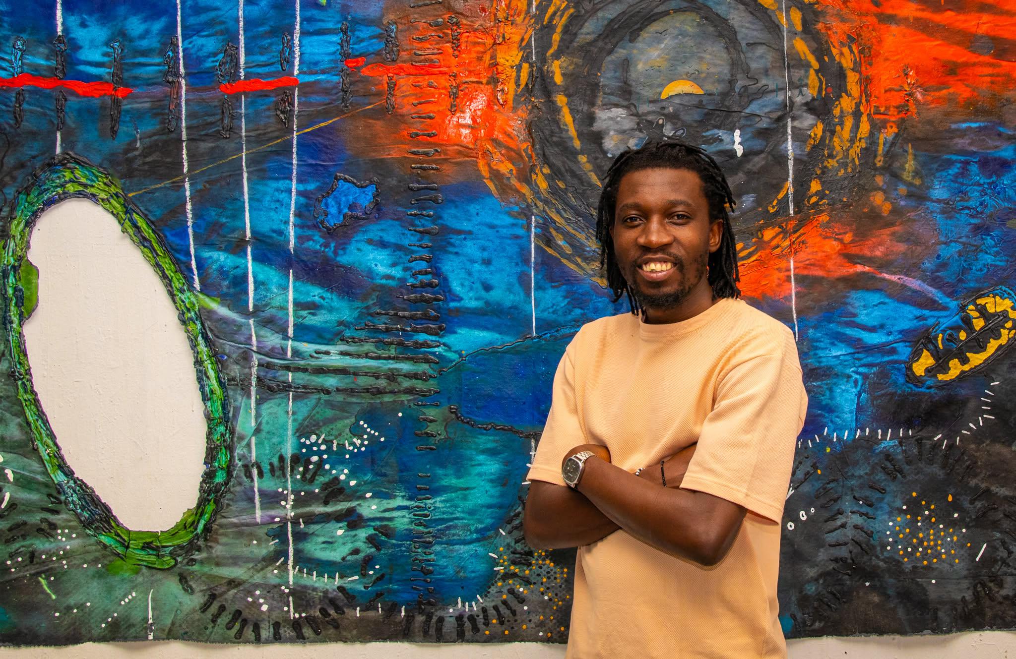 Person with long hair and a beard smiling in front of colorful abstract mural, arms crossed, wearing a beige shirt.